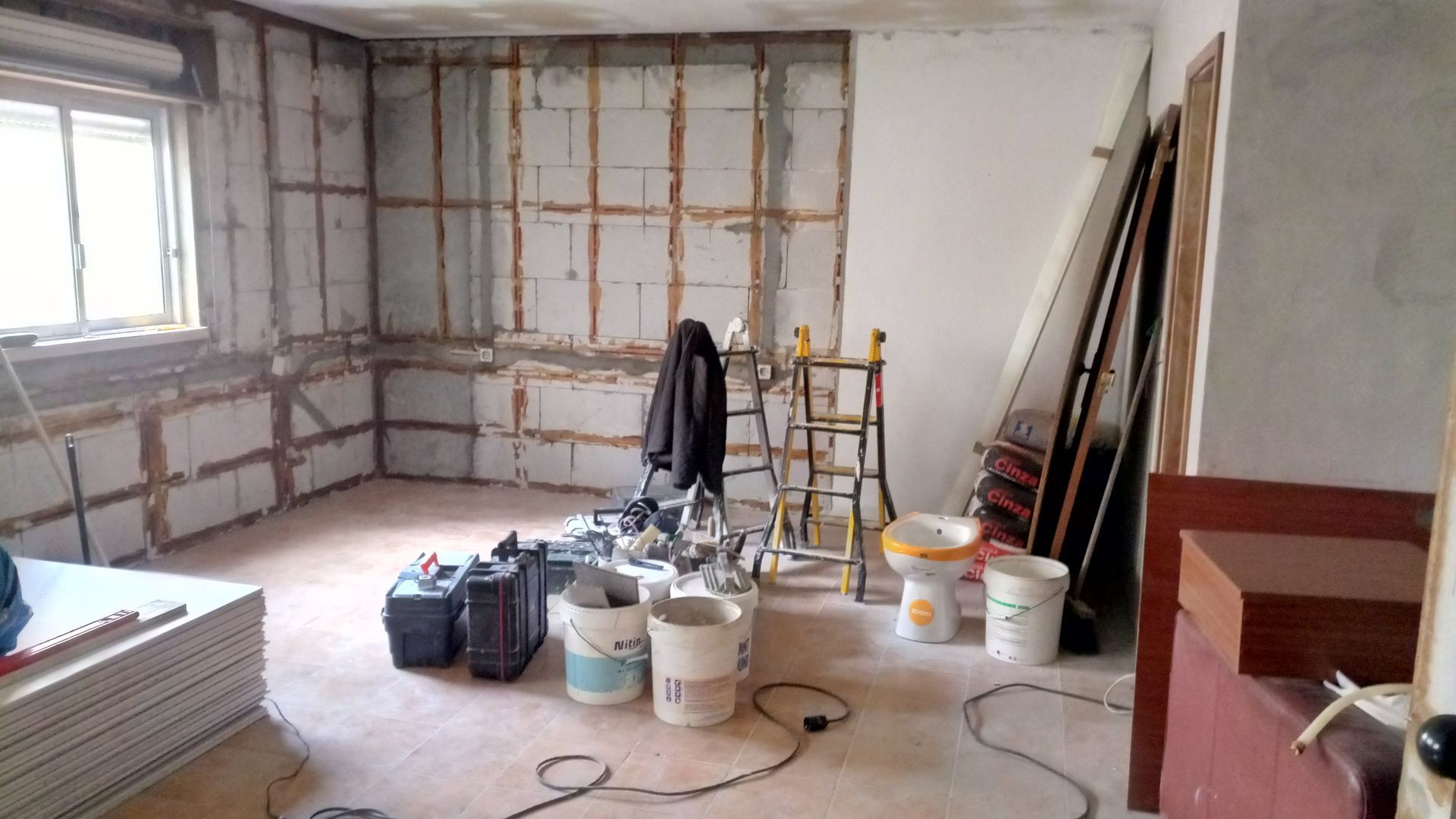 Quarto em reforma; paredes expostas, materiais de construção no chão, janela à esquerda, escada ao centro.