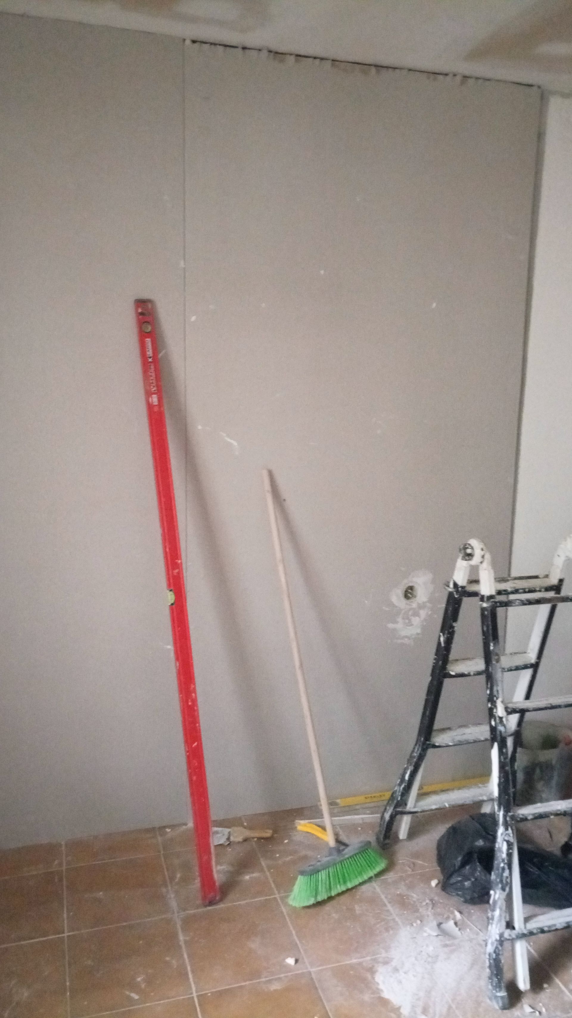 Canteiro de obras: Placas de gesso instaladas na parede, nível vermelho, vassoura, escada e entulho no chão.