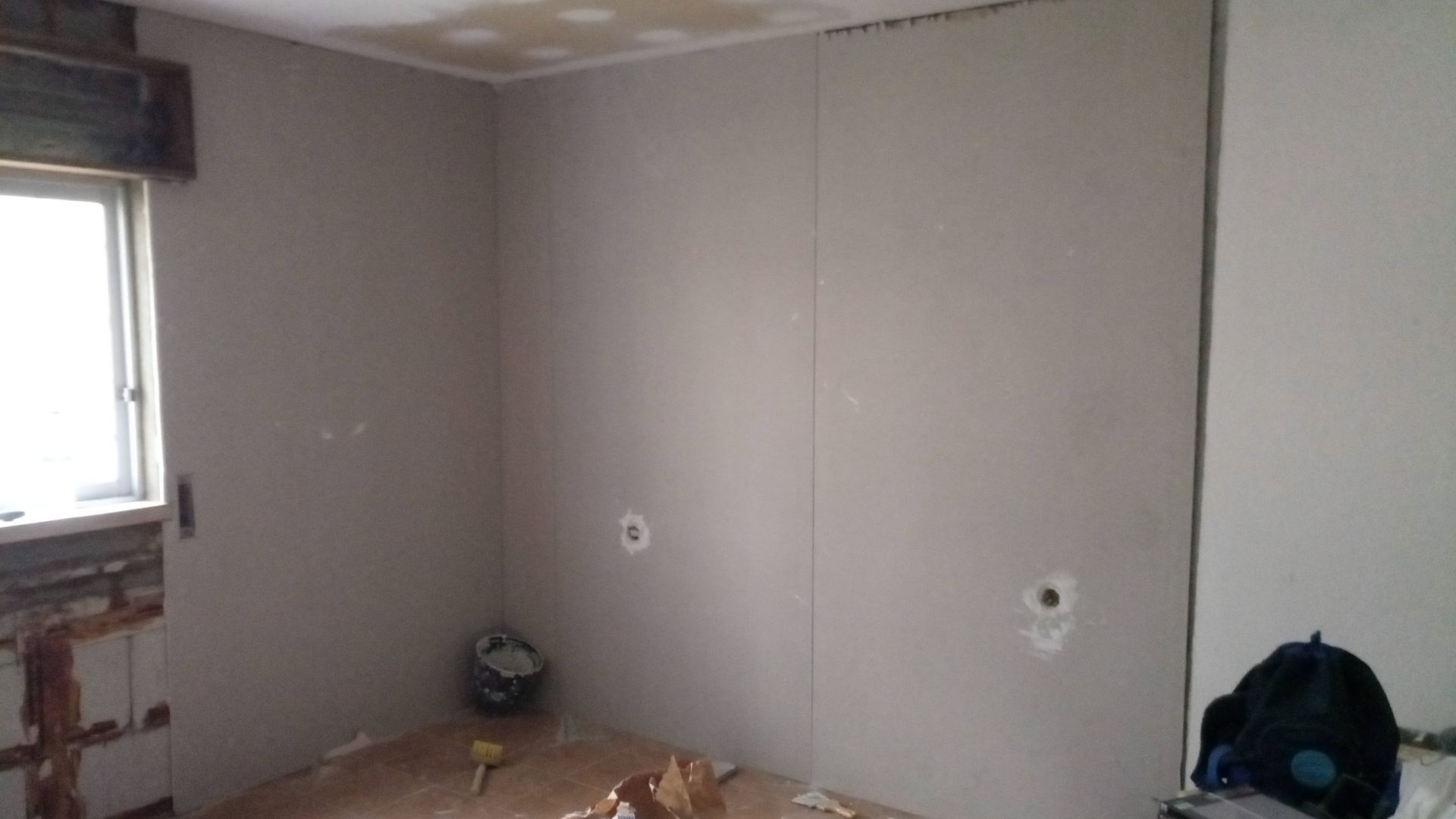 Quarto com drywall cinza instalado, inacabado, com furos para tomadas elétricas e janela à esquerda.