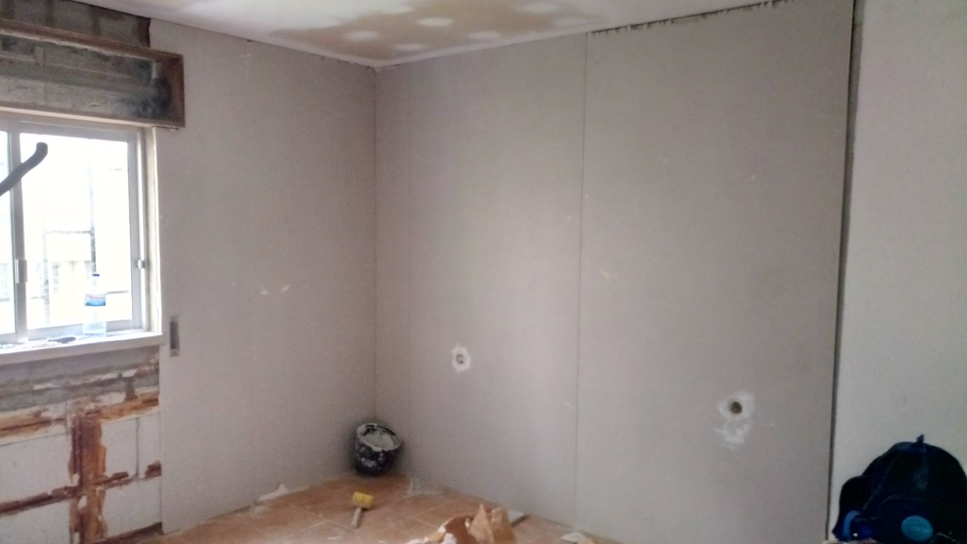 Interior do cômodo com paredes revestidas de gesso acartonado; janela exposta e madeira danificada.