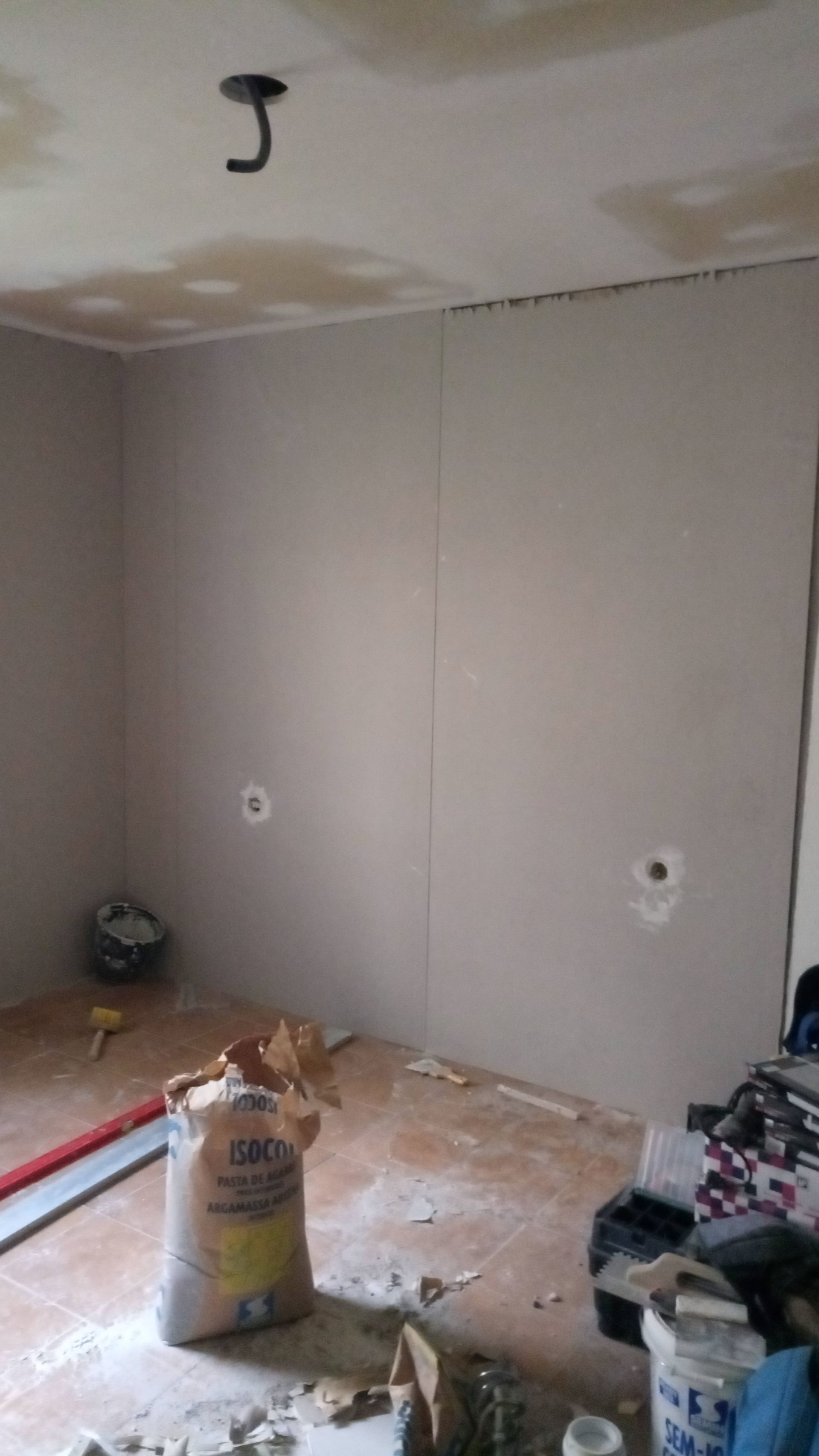 Quarto em reforma; paredes cinzentas, teto exposto, saco de material no chão, tomadas elétricas visíveis.