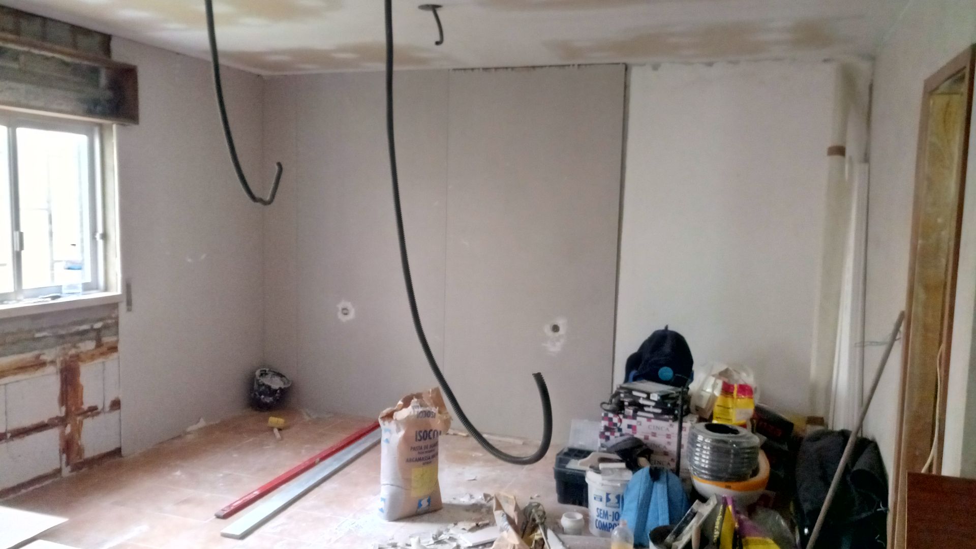 Sala em construção com fiação exposta, drywall e entulho no chão.