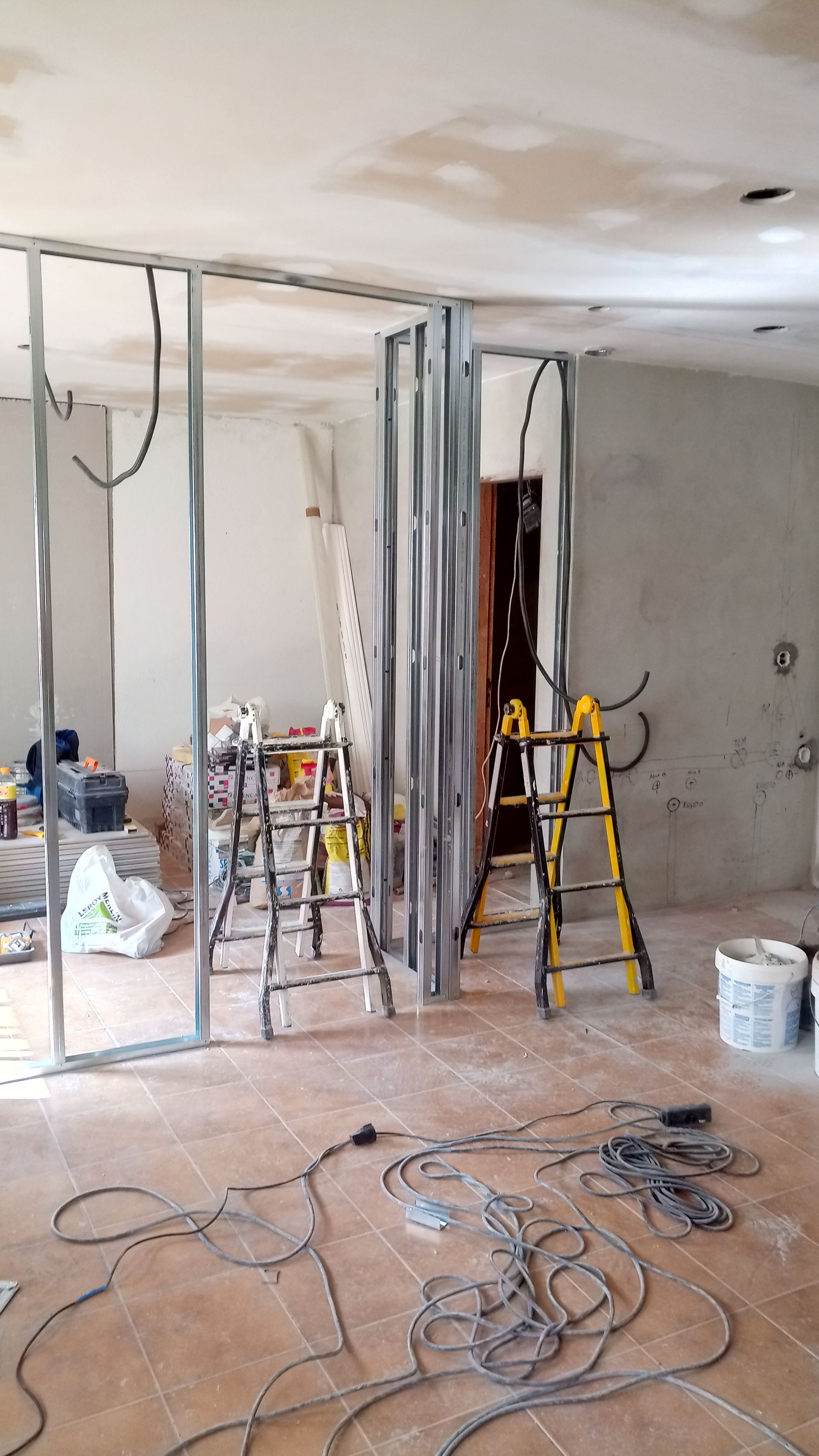 Canteiro de obras interno com perfis metálicos formando novas paredes; fiação elétrica, escadas e ferramentas presentes.