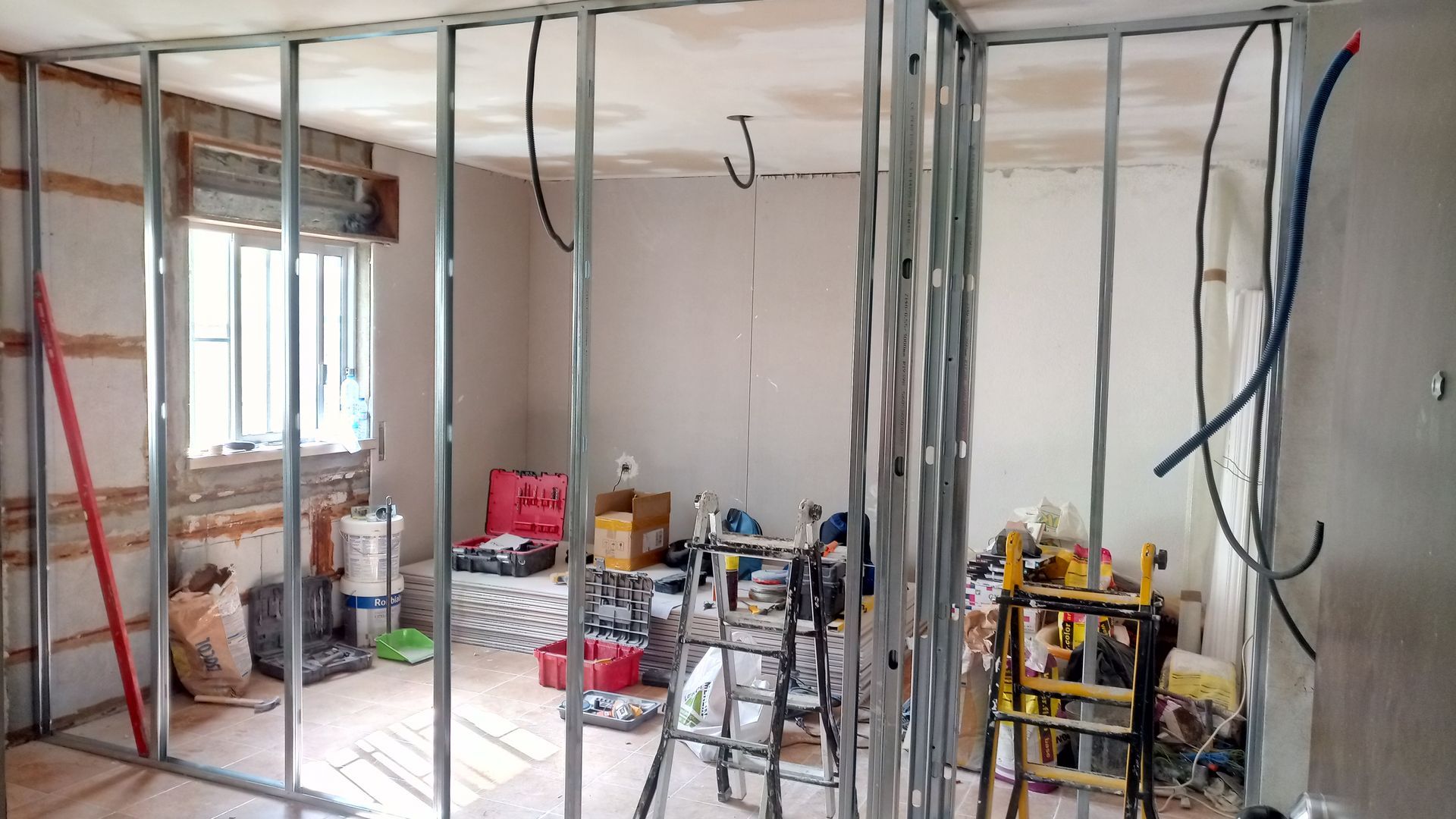 Interior de um cômodo com estrutura de metal em construção, fiação aparente e ferramentas.