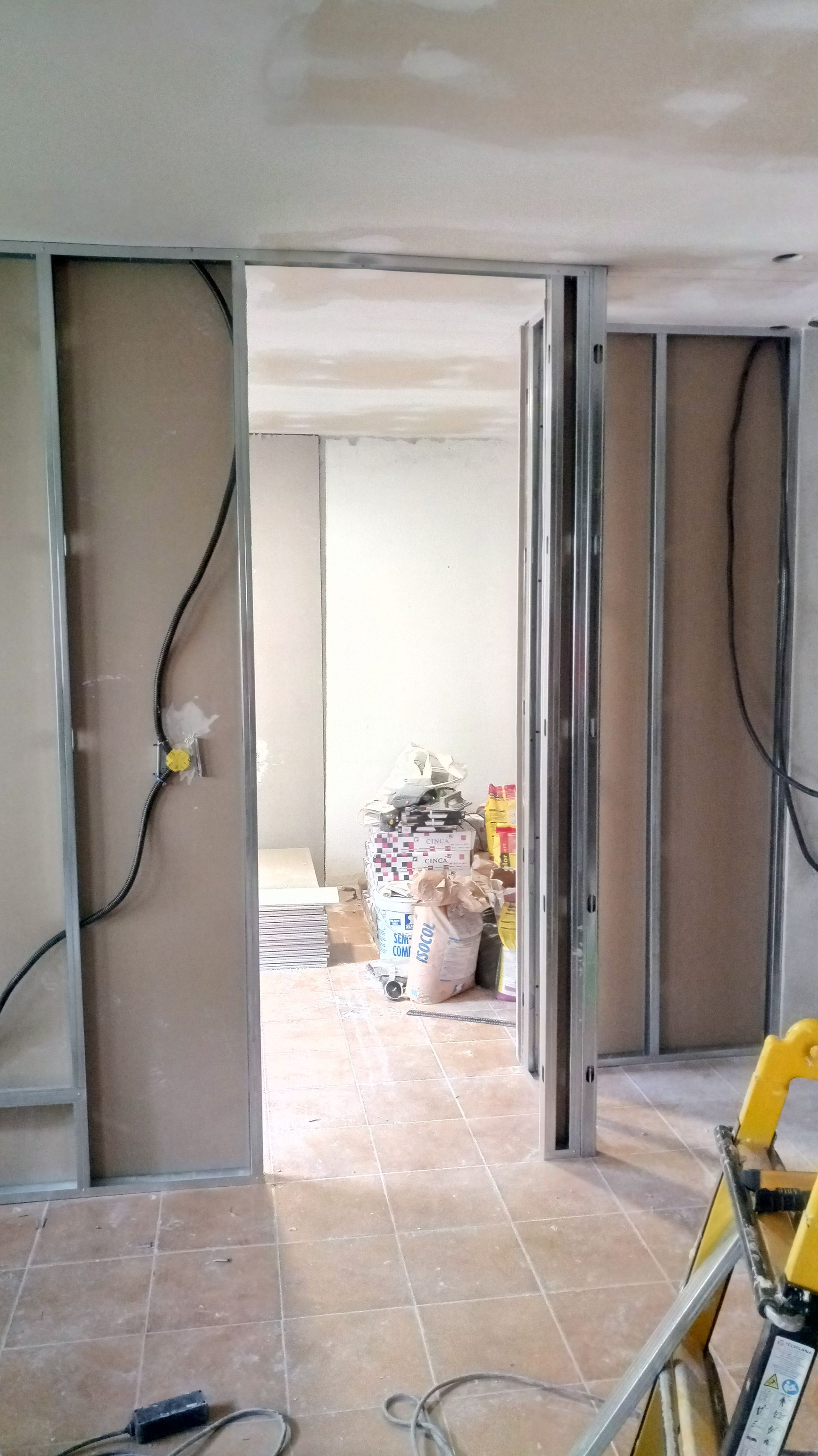 Parede interna em construção; painéis de drywall em molduras metálicas, porta aberta. Fios pretos e ferramentas visíveis.