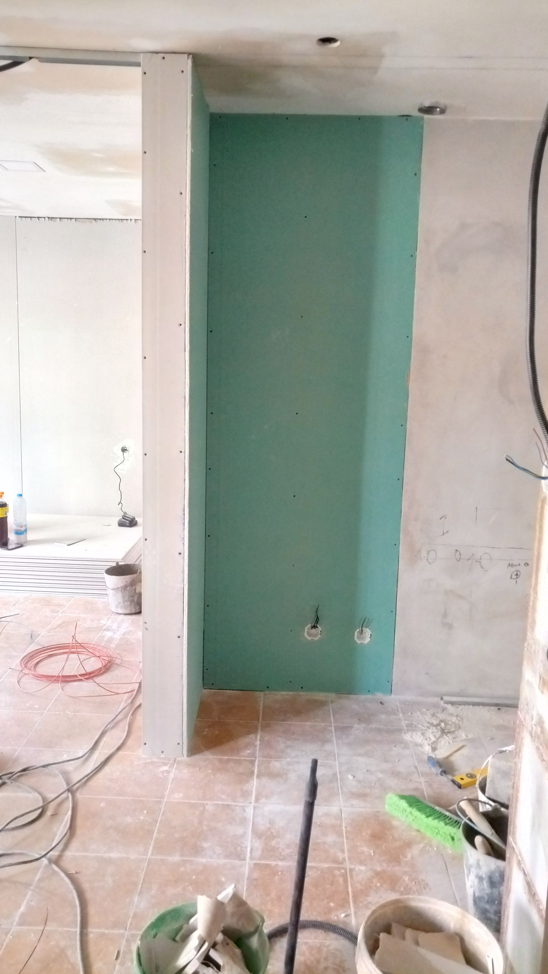 Vista interna de um cômodo em reforma; paredes de gesso acartonado verdes e sem acabamento; ferramentas e materiais espalhados pelo chão.