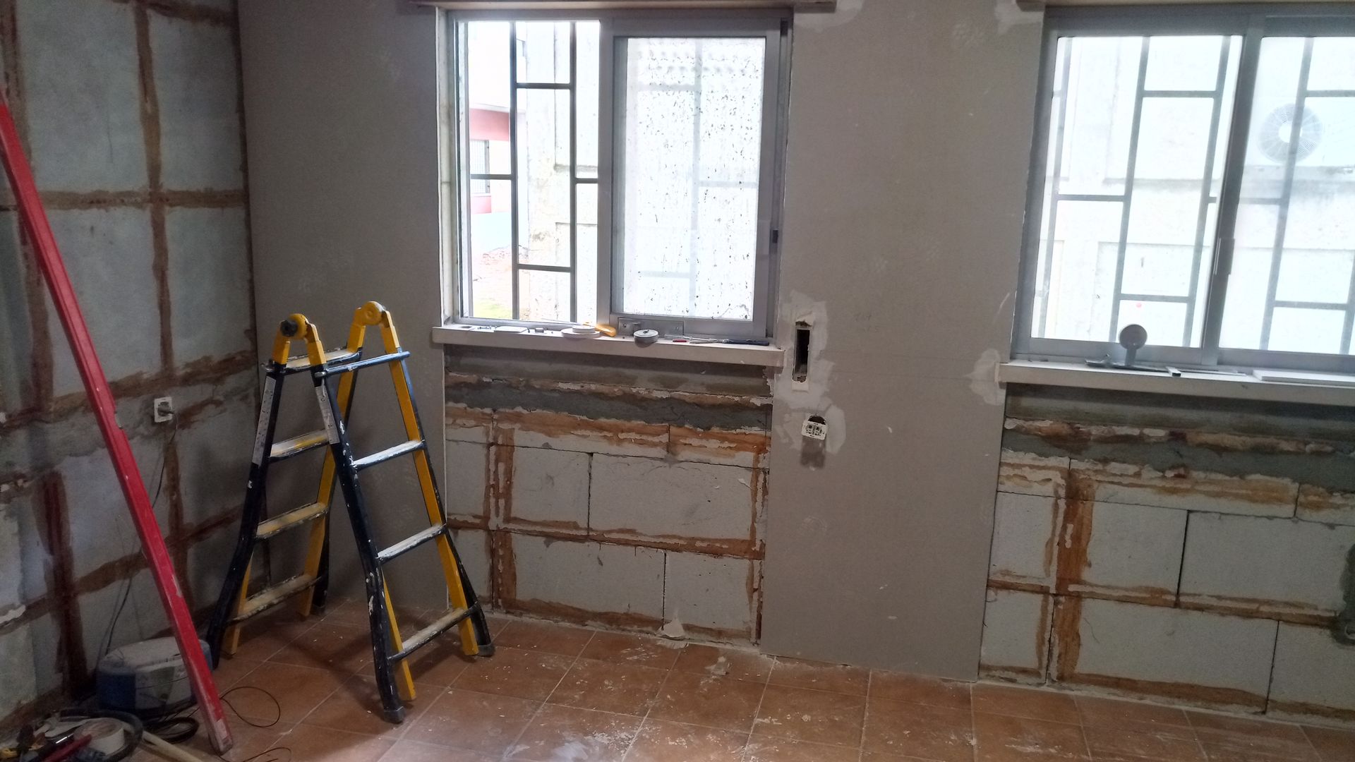 Interior de cômodo em construção com janelas, escada e estrutura de parede exposta.
