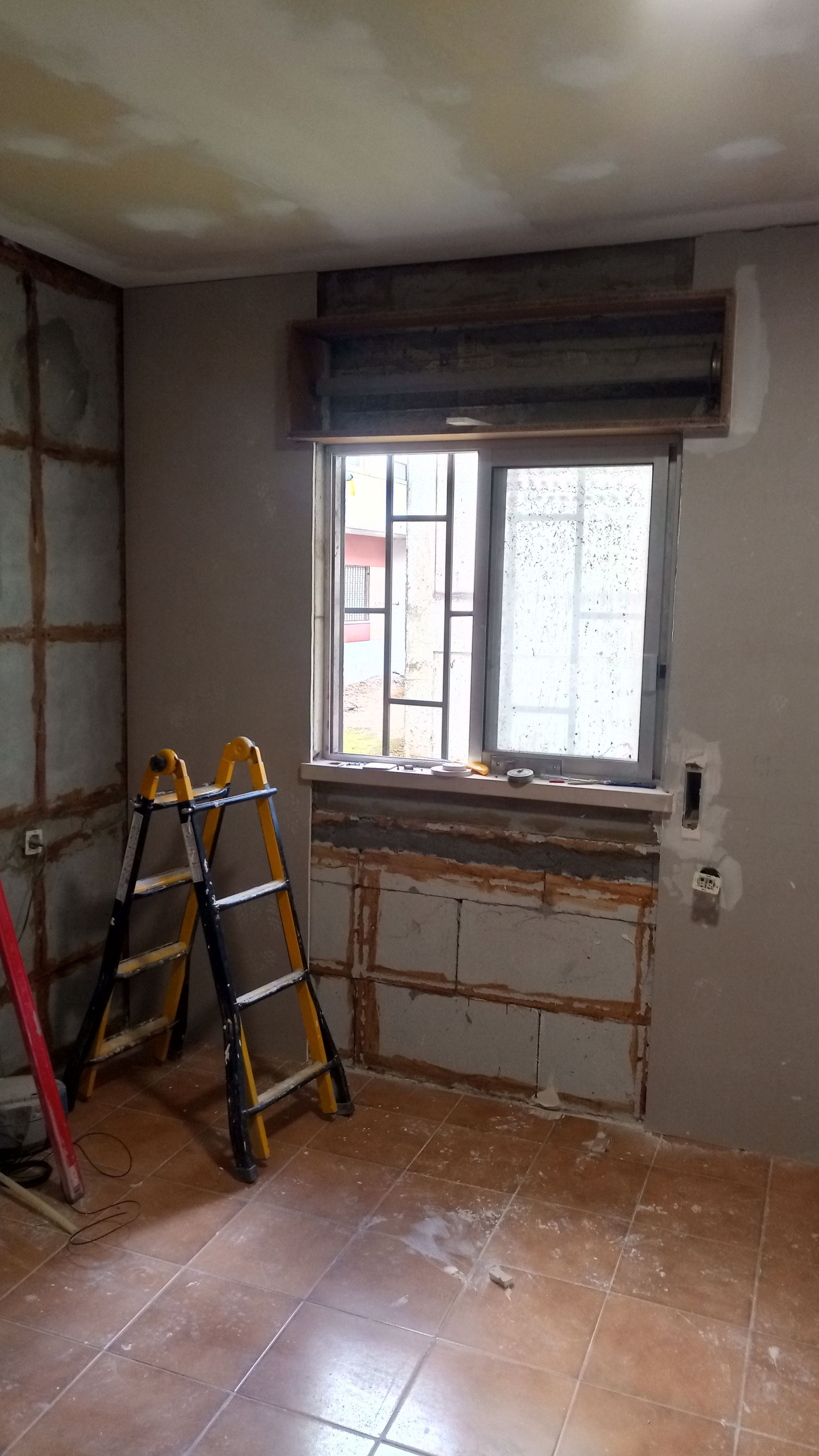 Quarto em reforma com escada, janela e painéis de parede danificados.