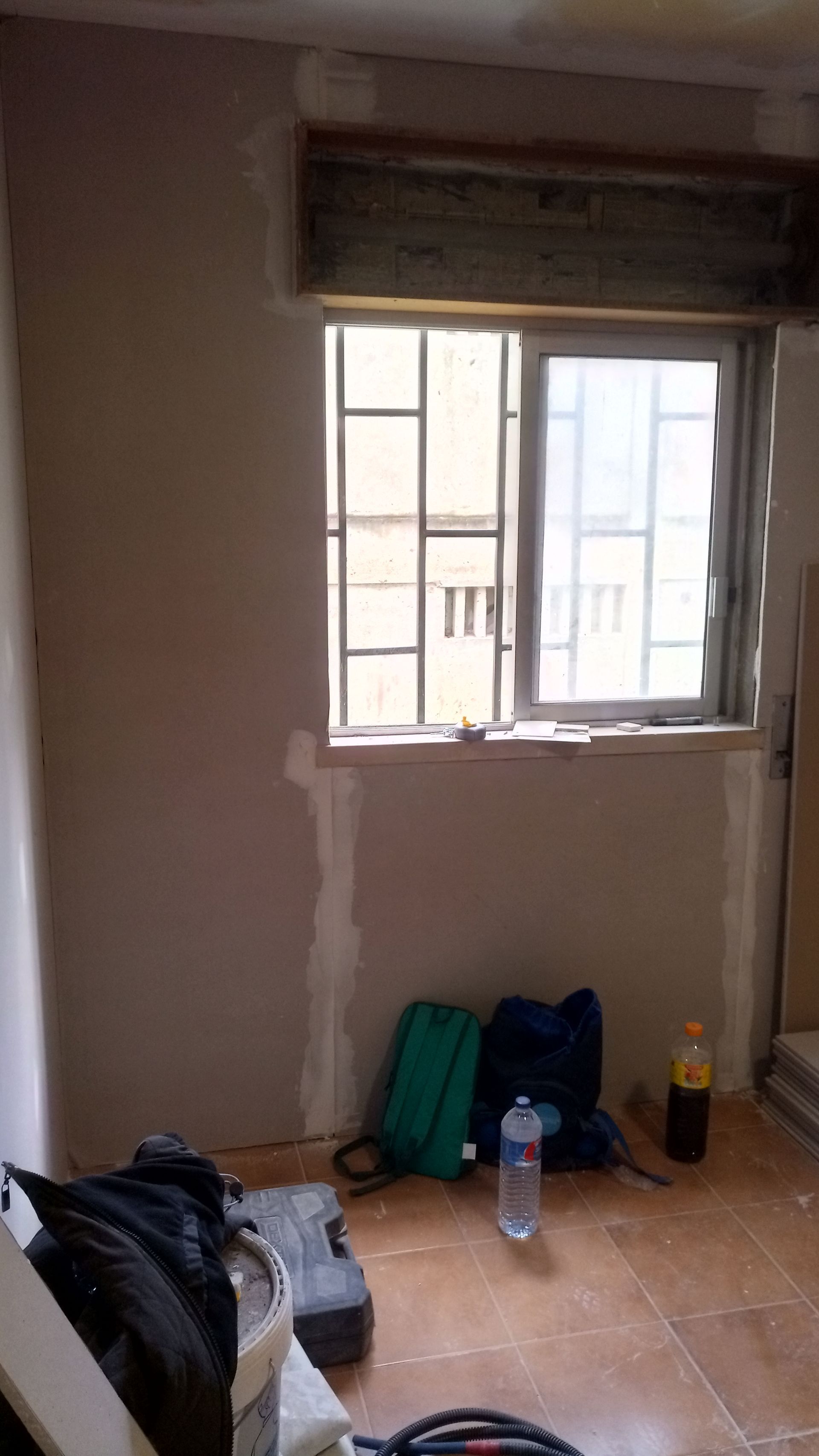 Interior de cômodo com janela, parede parcialmente reparada, materiais de construção no chão.