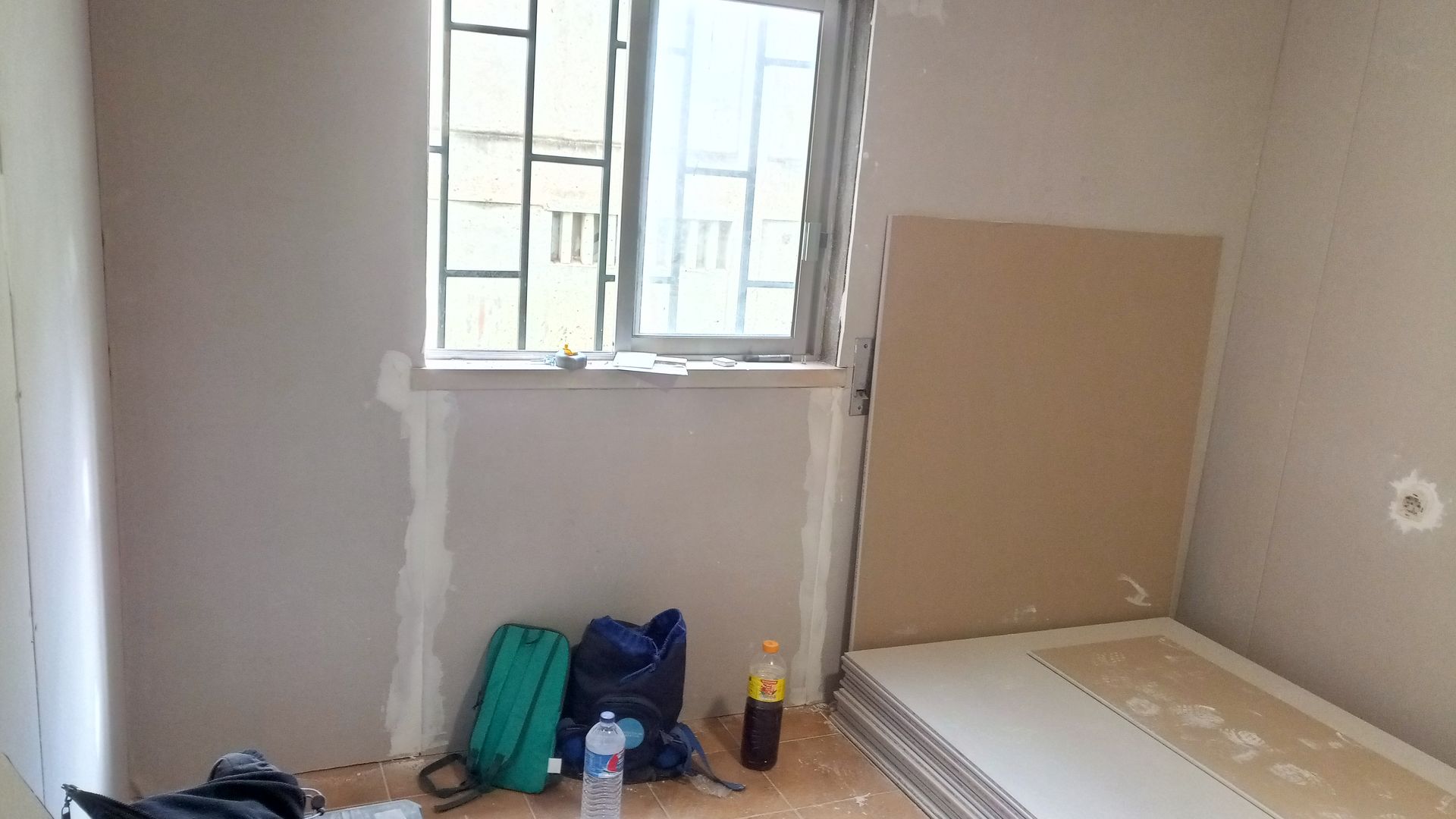 Um cômodo em construção com janela e parede de gesso. Sacolas e uma garrafa estão no chão.