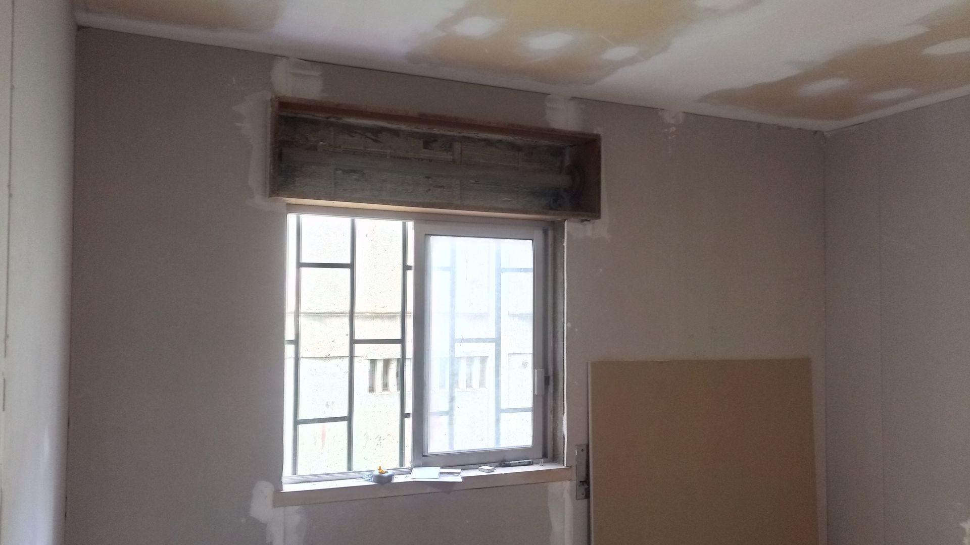 Interior do cômodo com janela, grades de segurança e persiana. Instalação de drywall em andamento.
