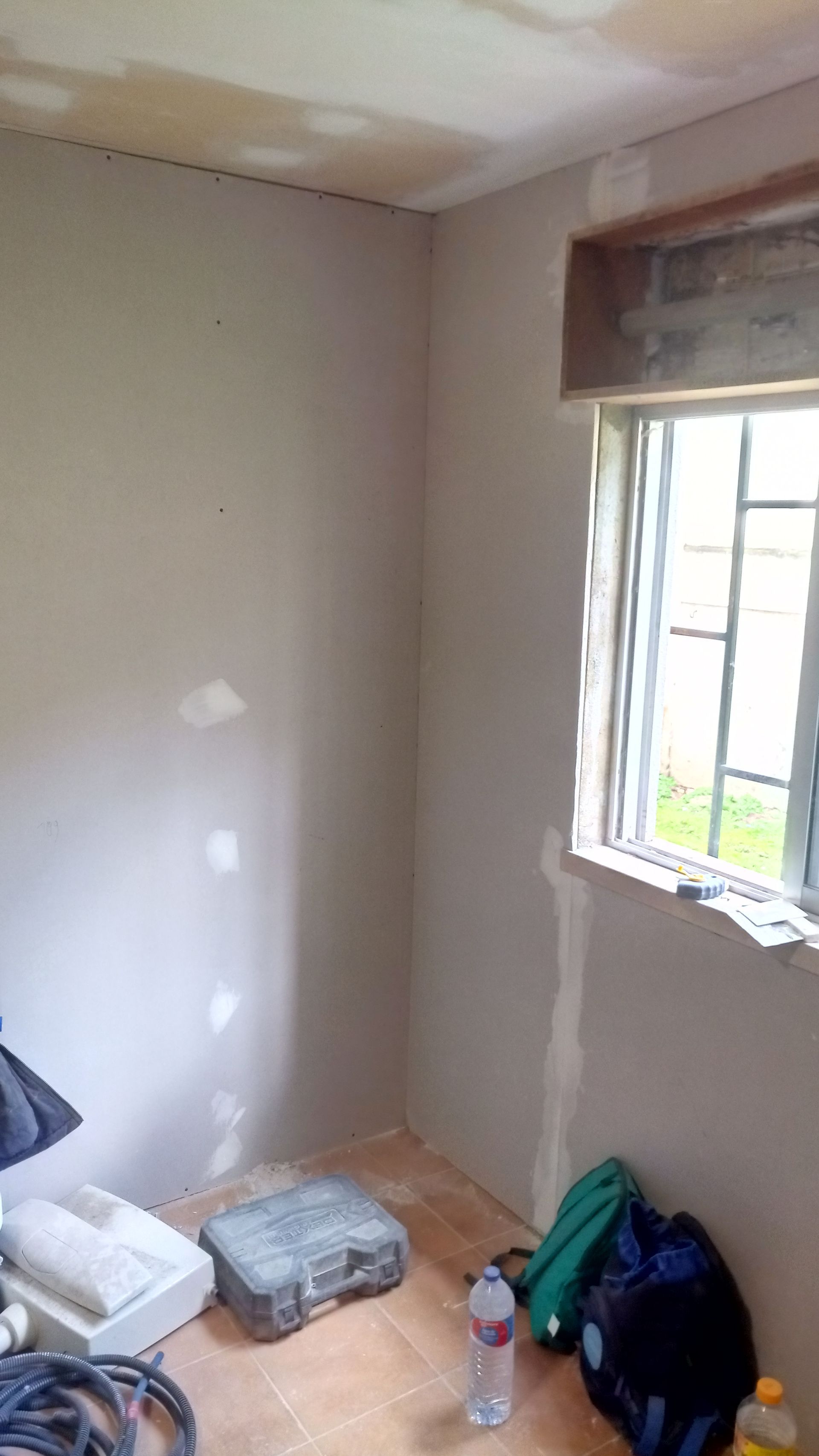 Canto de um cômodo em reforma; paredes cinzas, janela de madeira, ferramentas e materiais no chão.