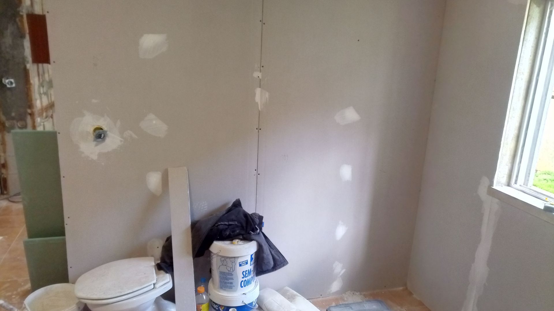Quarto interno em construção, com drywall remendado, vaso sanitário, janela e materiais de construção.