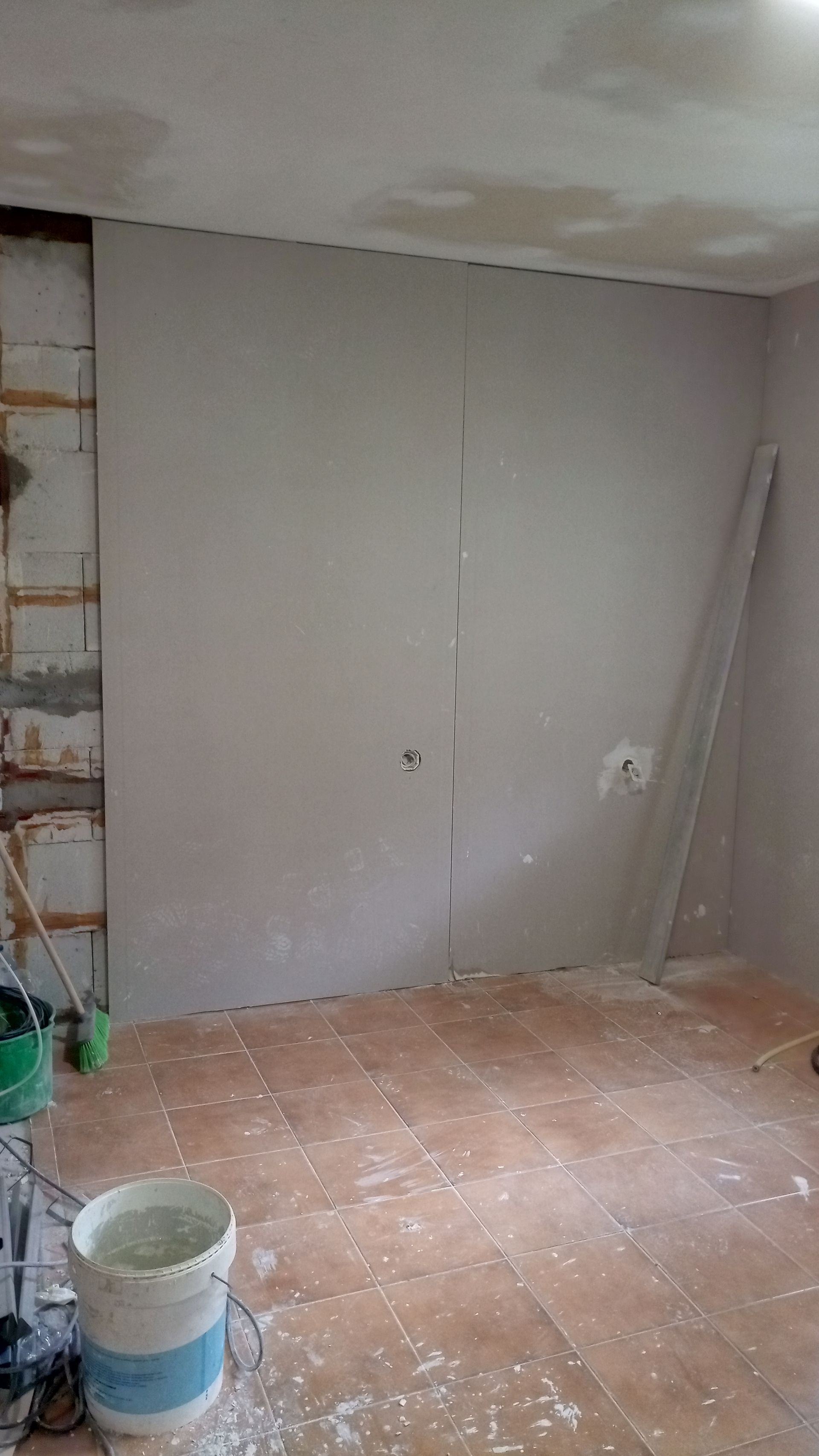 Cômodo em construção, com drywall em uma parede. Bloco de concreto, balde de tinta e piso de madeira visíveis.