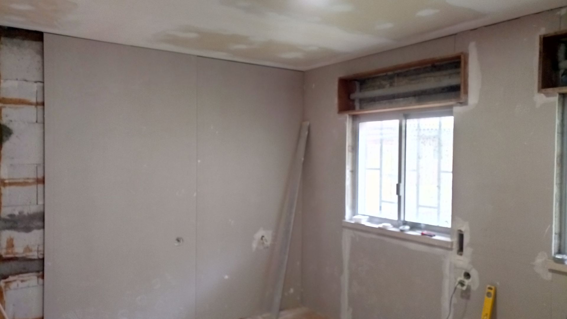 Quarto em reforma com paredes de gesso cartonado, uma janela e teto inacabado.