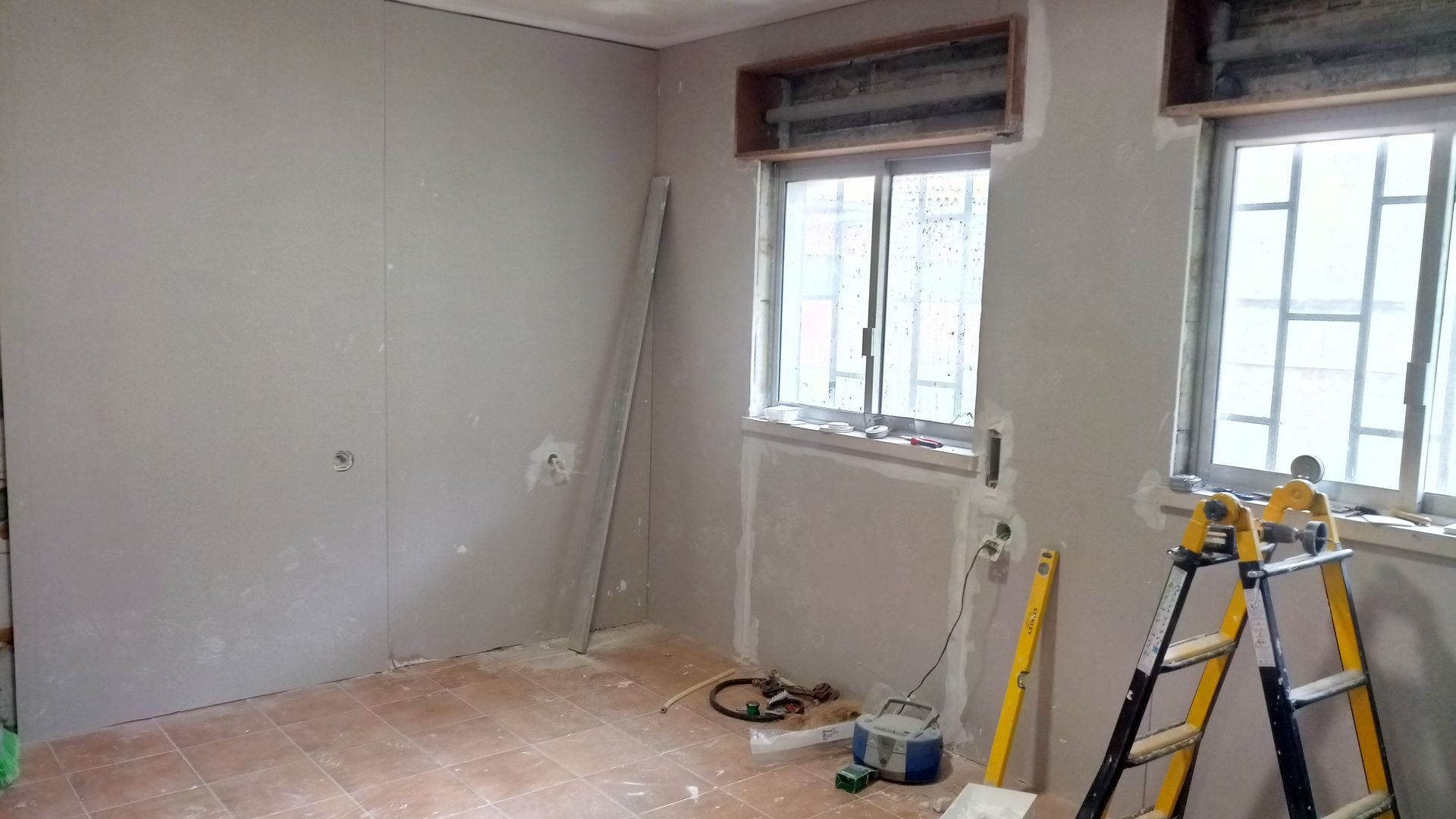 Cômodo em construção; drywall, duas janelas, escada, ferramentas, entulho no chão, paredes cinza-claro.