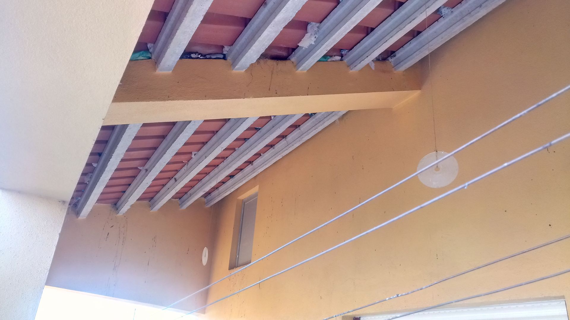 Parede externa amarela com vigas aparentes no teto e telhas de terracota. Varal de roupas branco atravessa a parede.