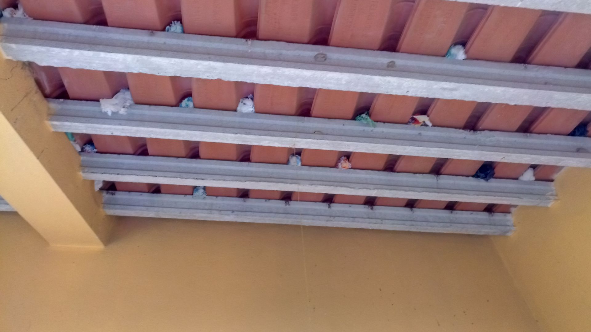 Vista de um teto com vigas de concreto e telhas de barro vermelho, sustentado por uma parede amarela.