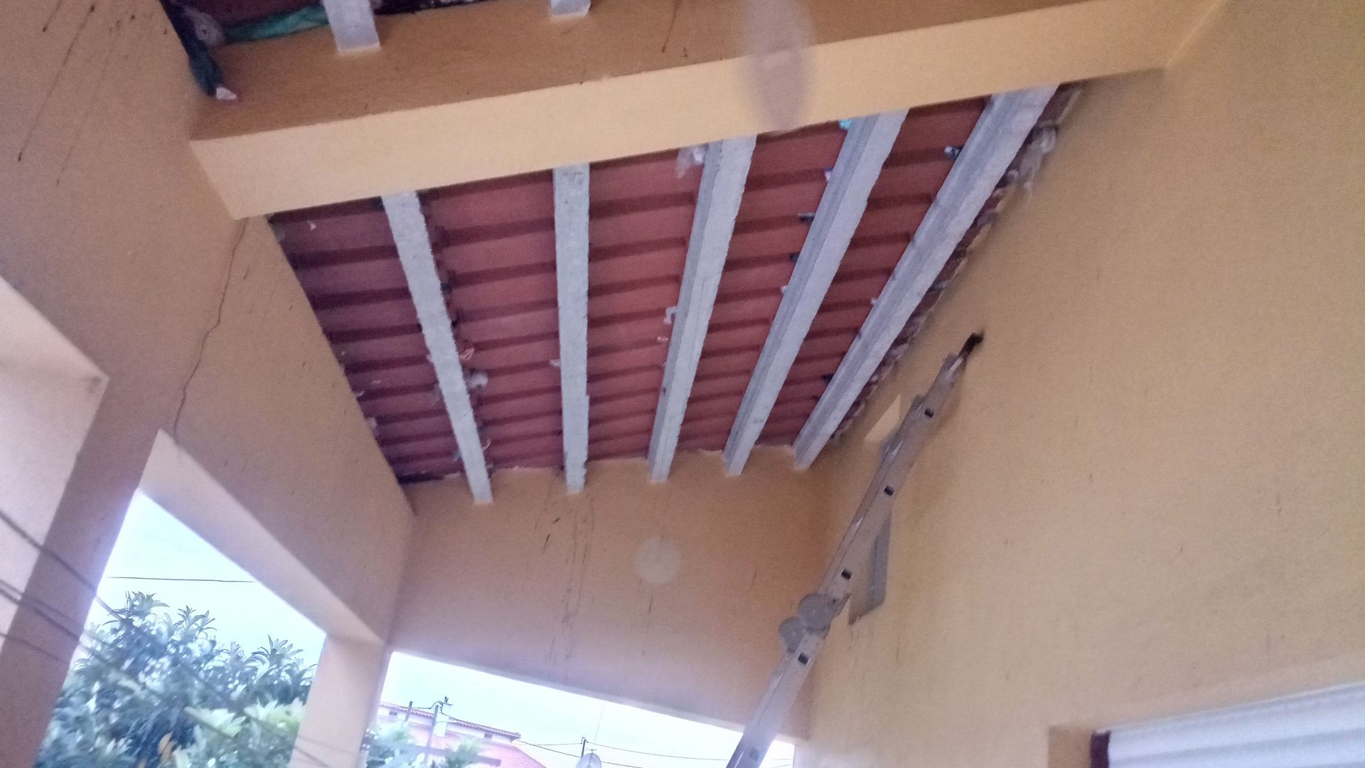 Vista externa do teto de uma varanda em construção; telhas laranja, vigas brancas, escada encostada na parede amarela.
