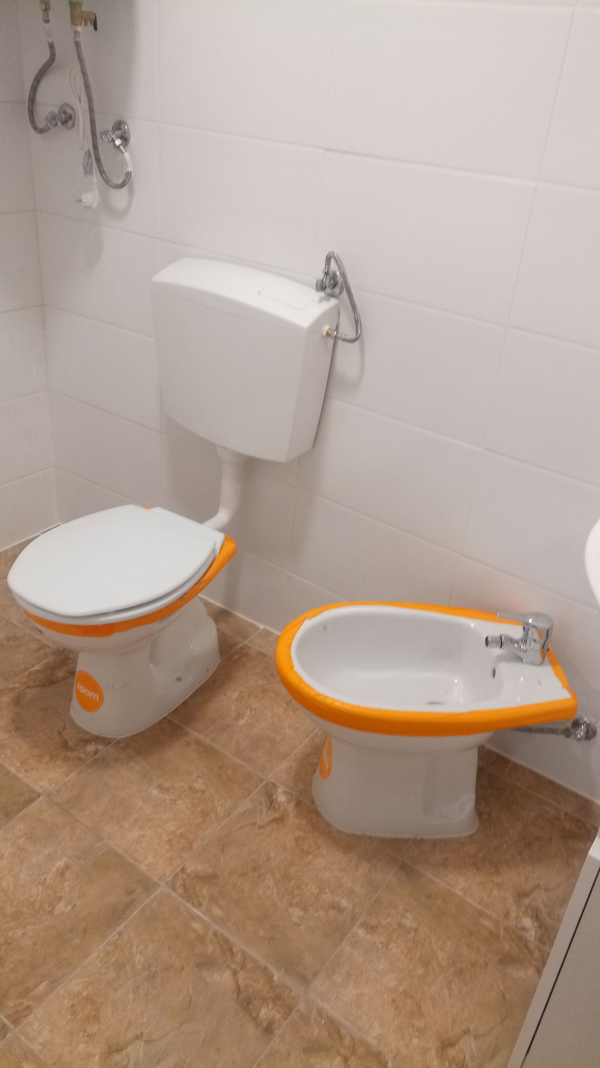 Vaso sanitário e bidê em um banheiro. Louças sanitárias brancas com detalhes em laranja sobre piso de azulejo.