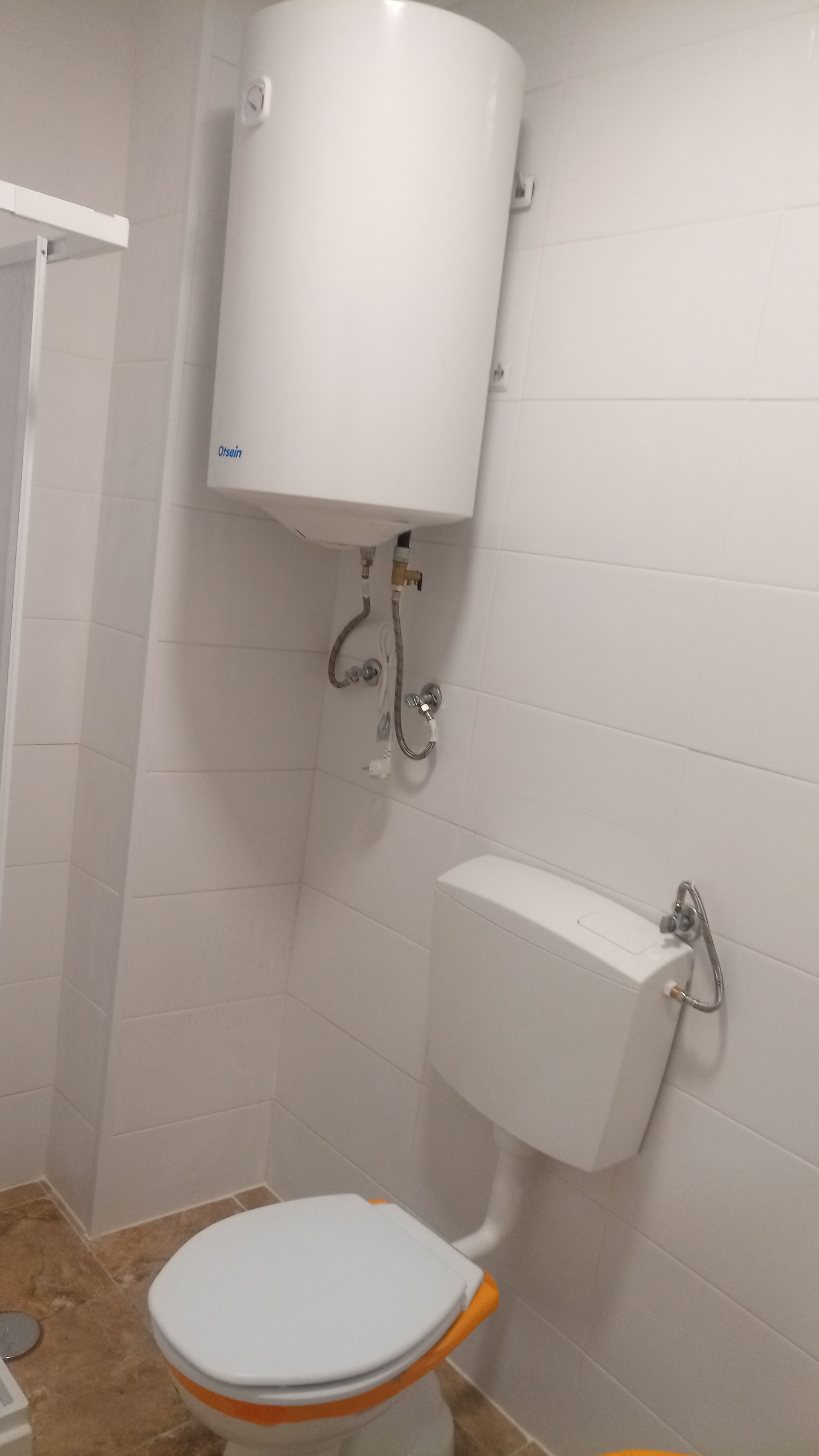 Aquecedor de água branco acima do vaso sanitário em um banheiro com paredes revestidas de azulejos brancos e piso bege.