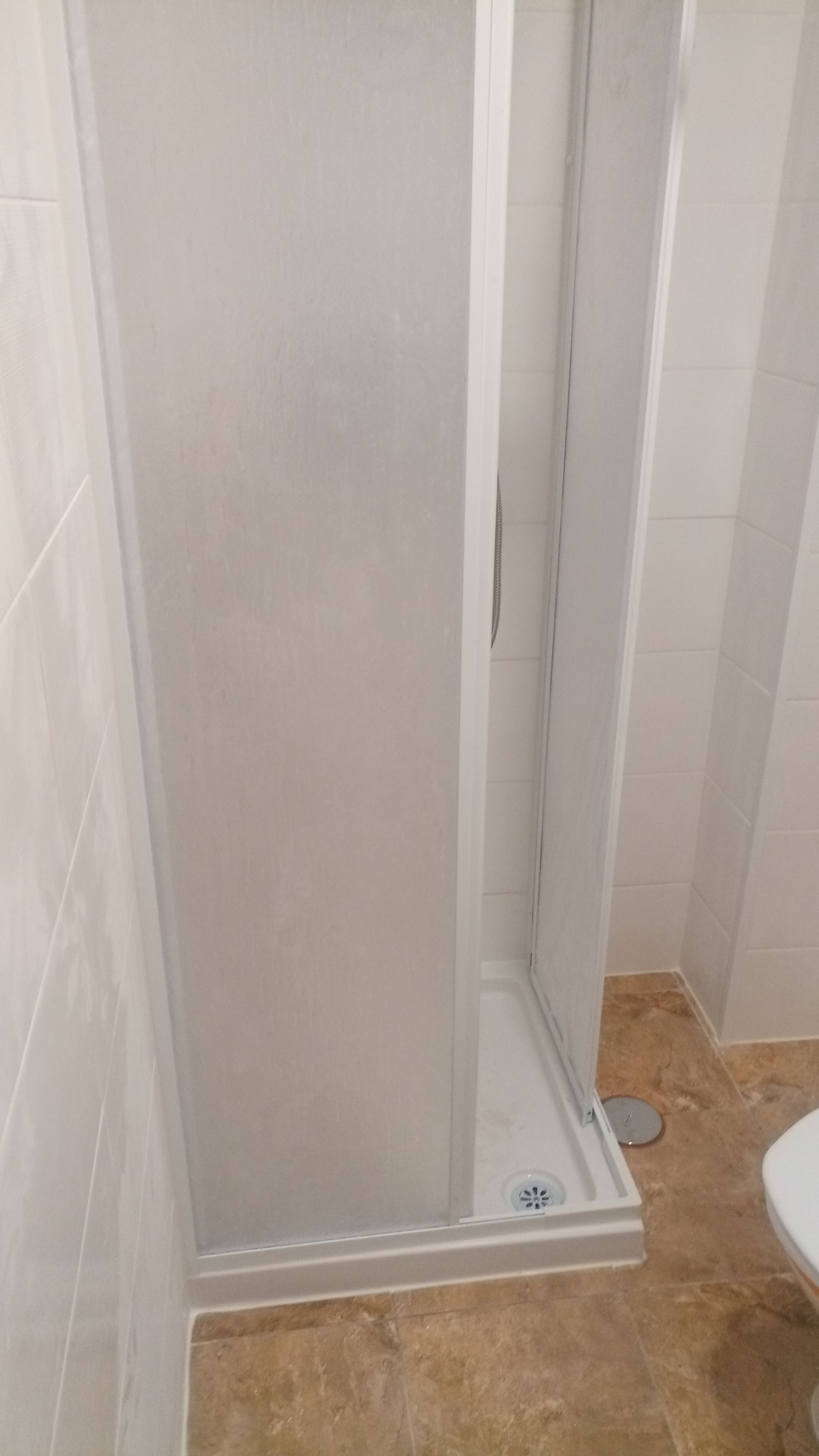 Cabina de duche branca com porta de correr, localizada entre paredes revestidas a azulejos e um piso bege.