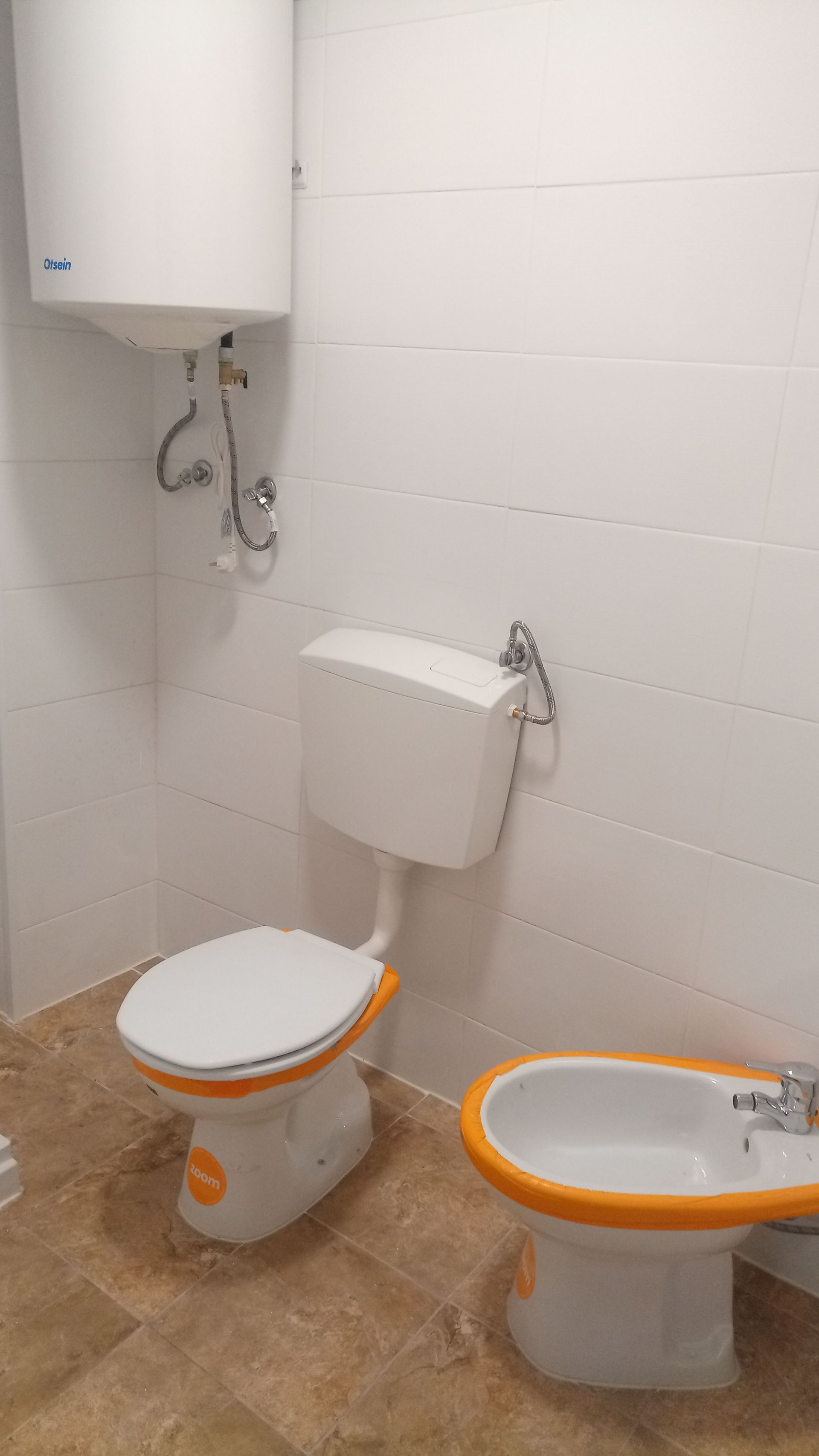 Banheiro com paredes revestidas de azulejos brancos, vaso sanitário, bidê e aquecedor de água; detalhes em laranja.