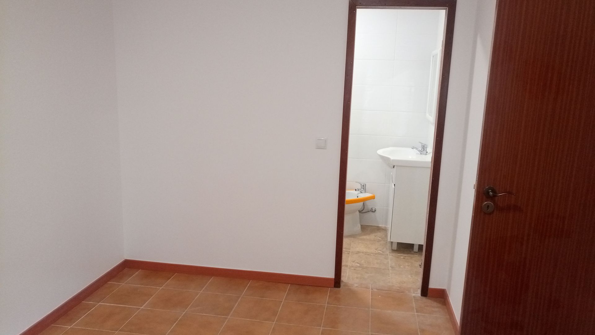 Quarto vazio com piso de azulejo e porta que dá acesso a um pequeno banheiro com pia e vaso sanitário.