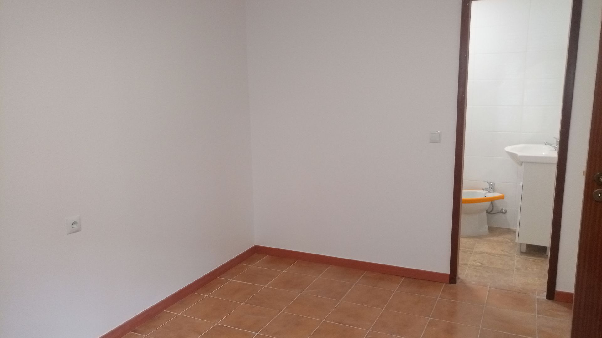 Quarto vazio com paredes brancas, piso de azulejo marrom e porta que dá acesso a um banheiro com pia e vaso sanitário.