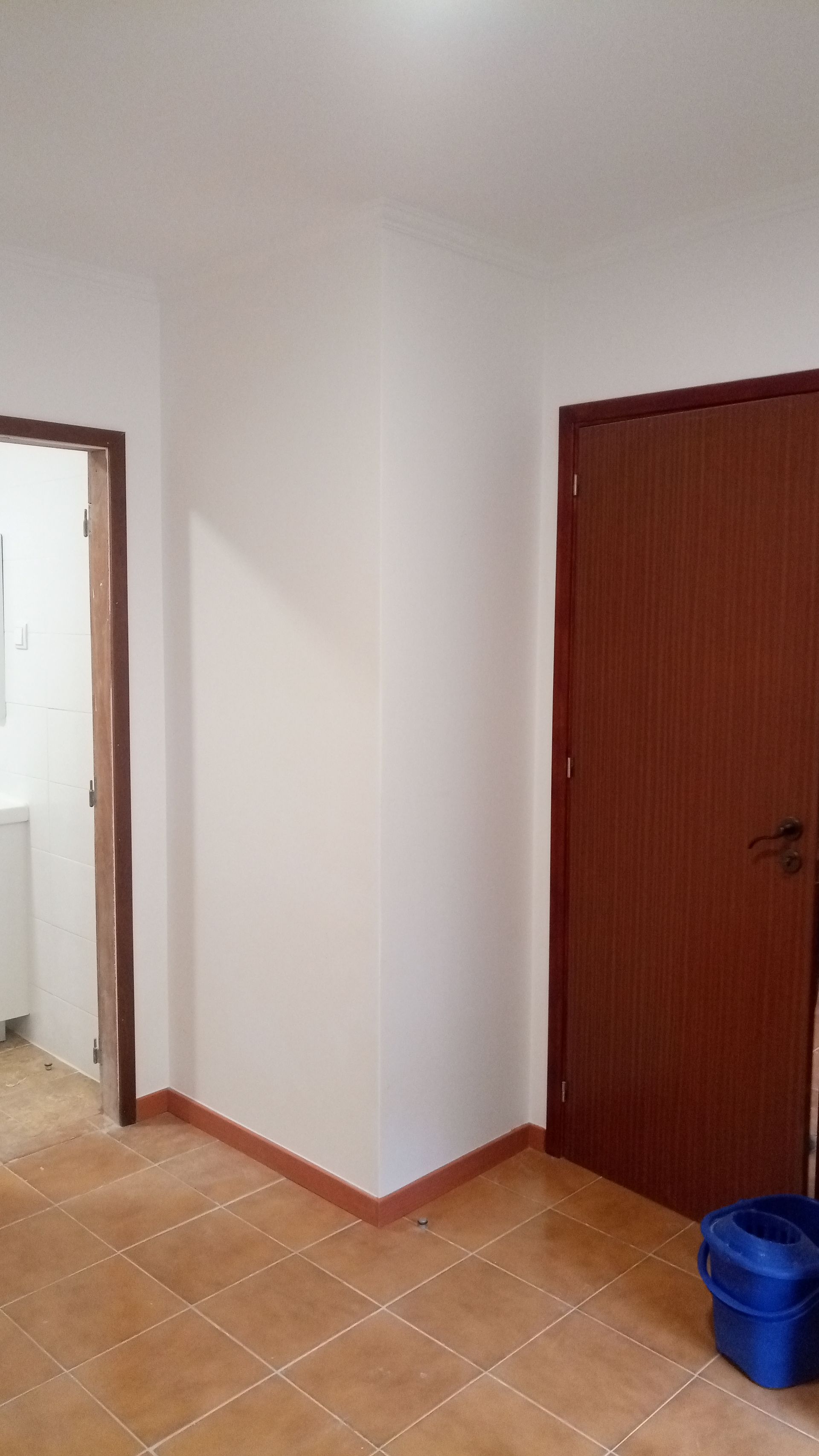 Canto interno com portas e piso de ladrilho cor terracota. Batentes de porta marrons, paredes brancas, balde azul.
