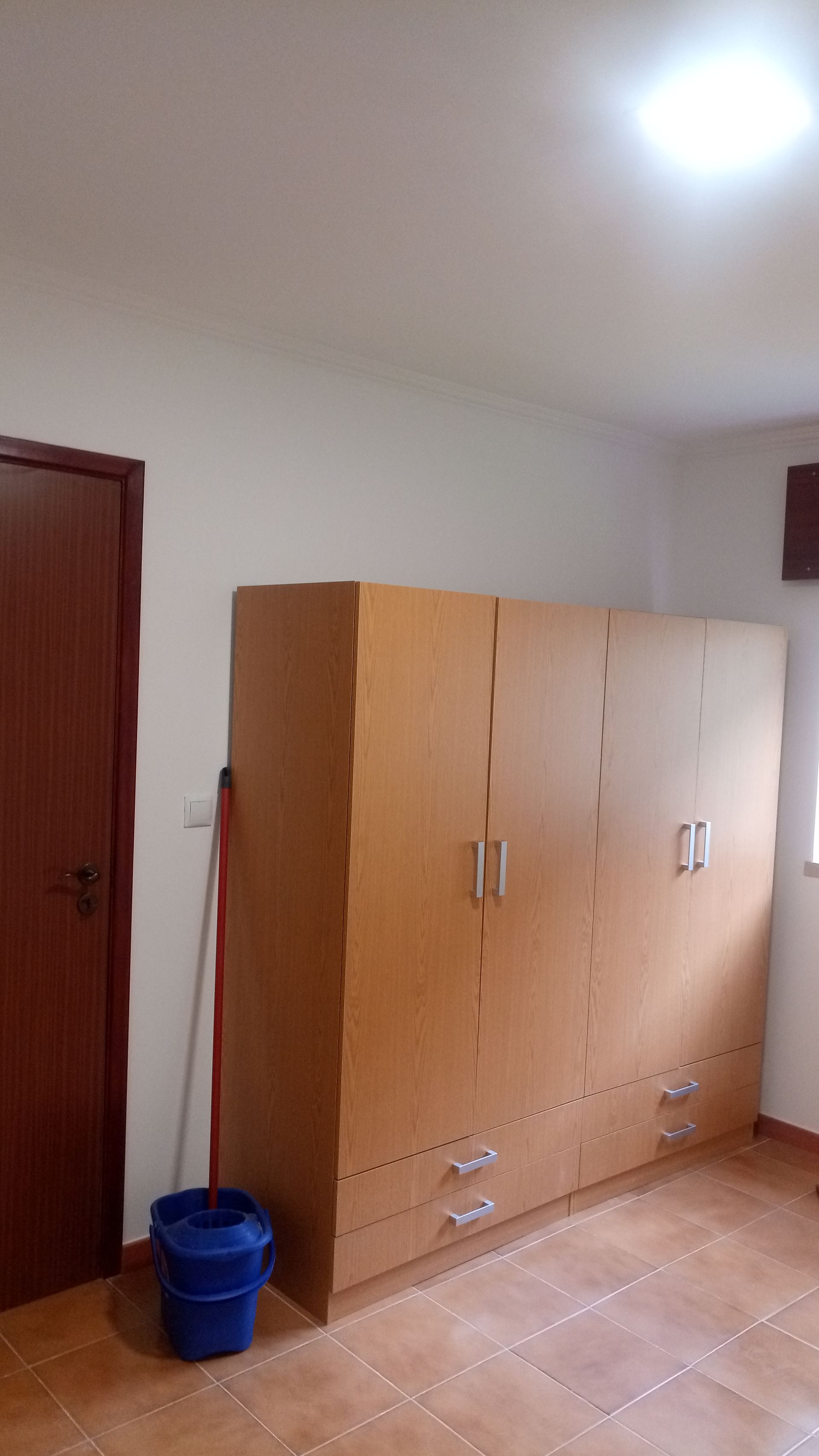 Quarto com um grande guarda-roupa de madeira e uma porta marrom fechada. Um balde azul está encostado no guarda-roupa.