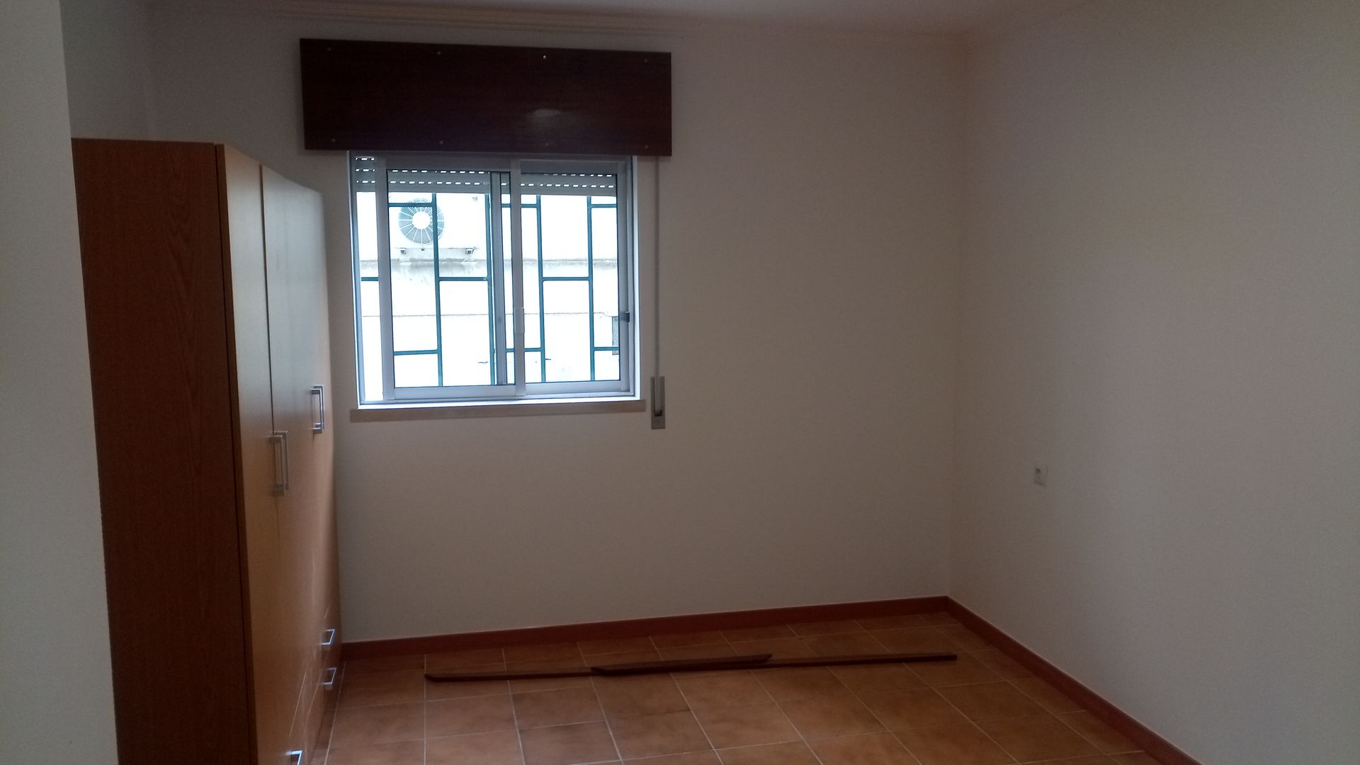 Quarto vazio com janela, guarda-roupa e piso de azulejo marrom. Paredes bege e persiana marrom.