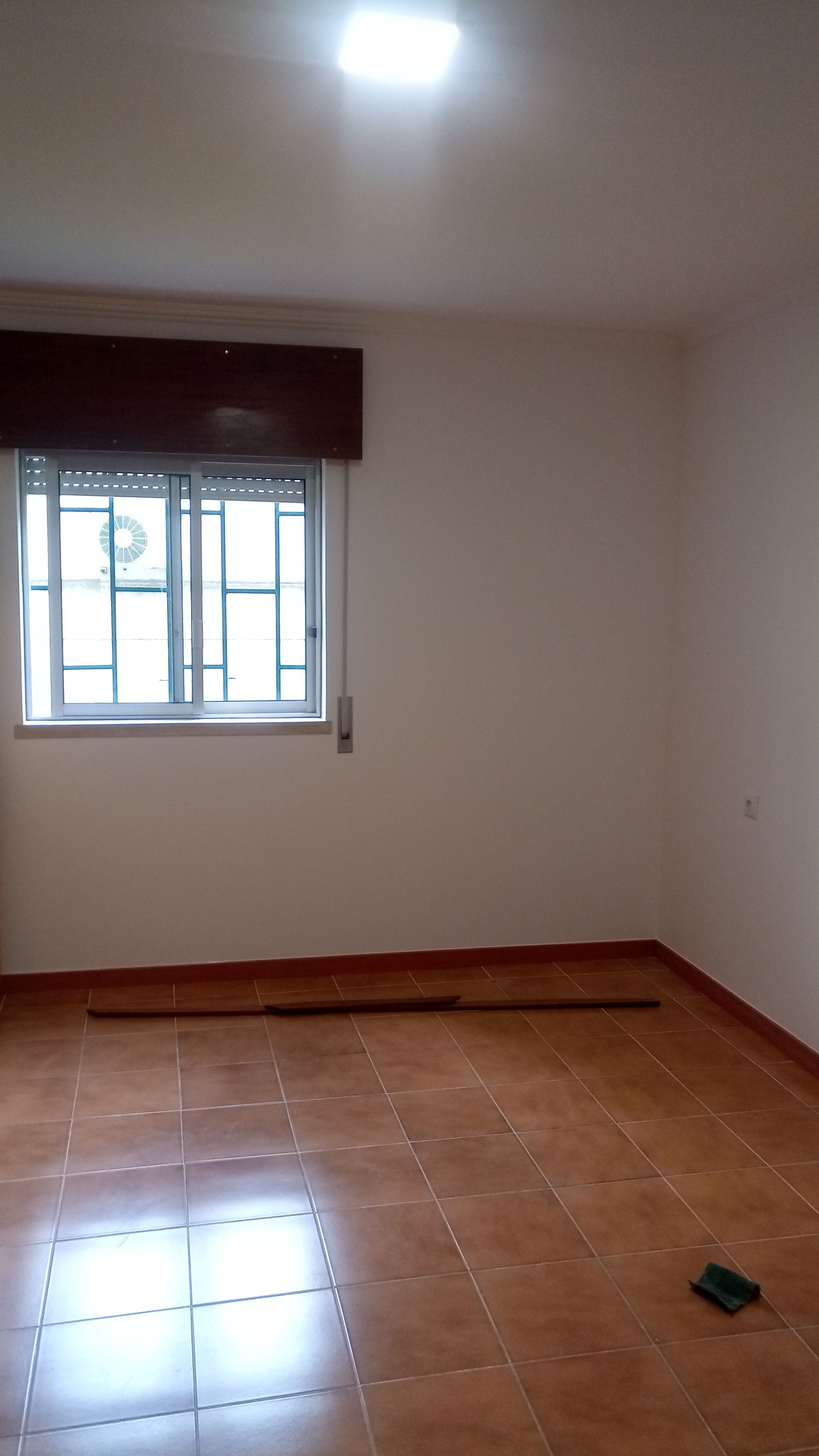 Quarto vazio com janela, piso de azulejo marrom e luminária branca no teto.