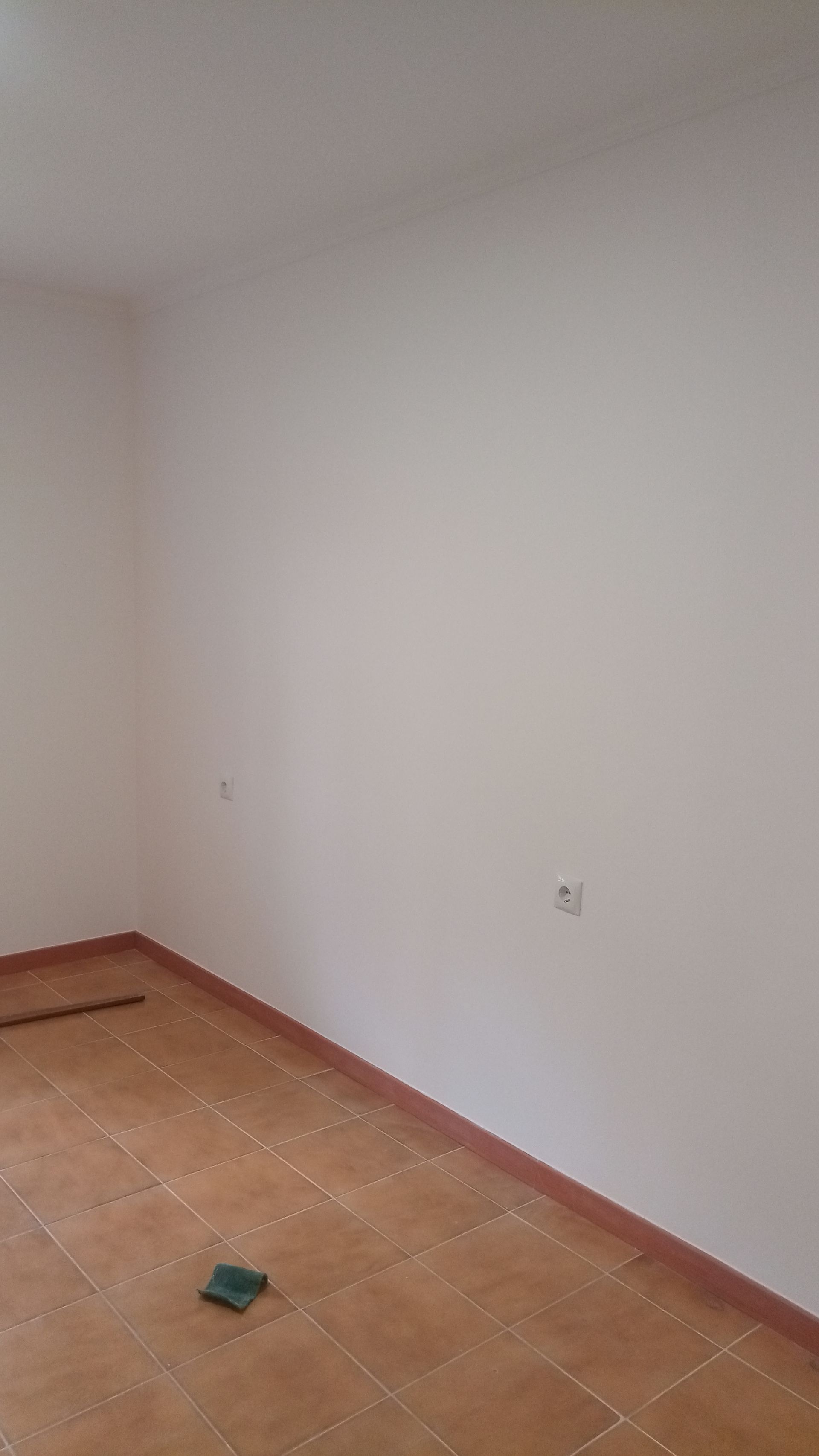 Quarto vazio com paredes bege e piso de ladrilho terracota; um interruptor de luz é visível.