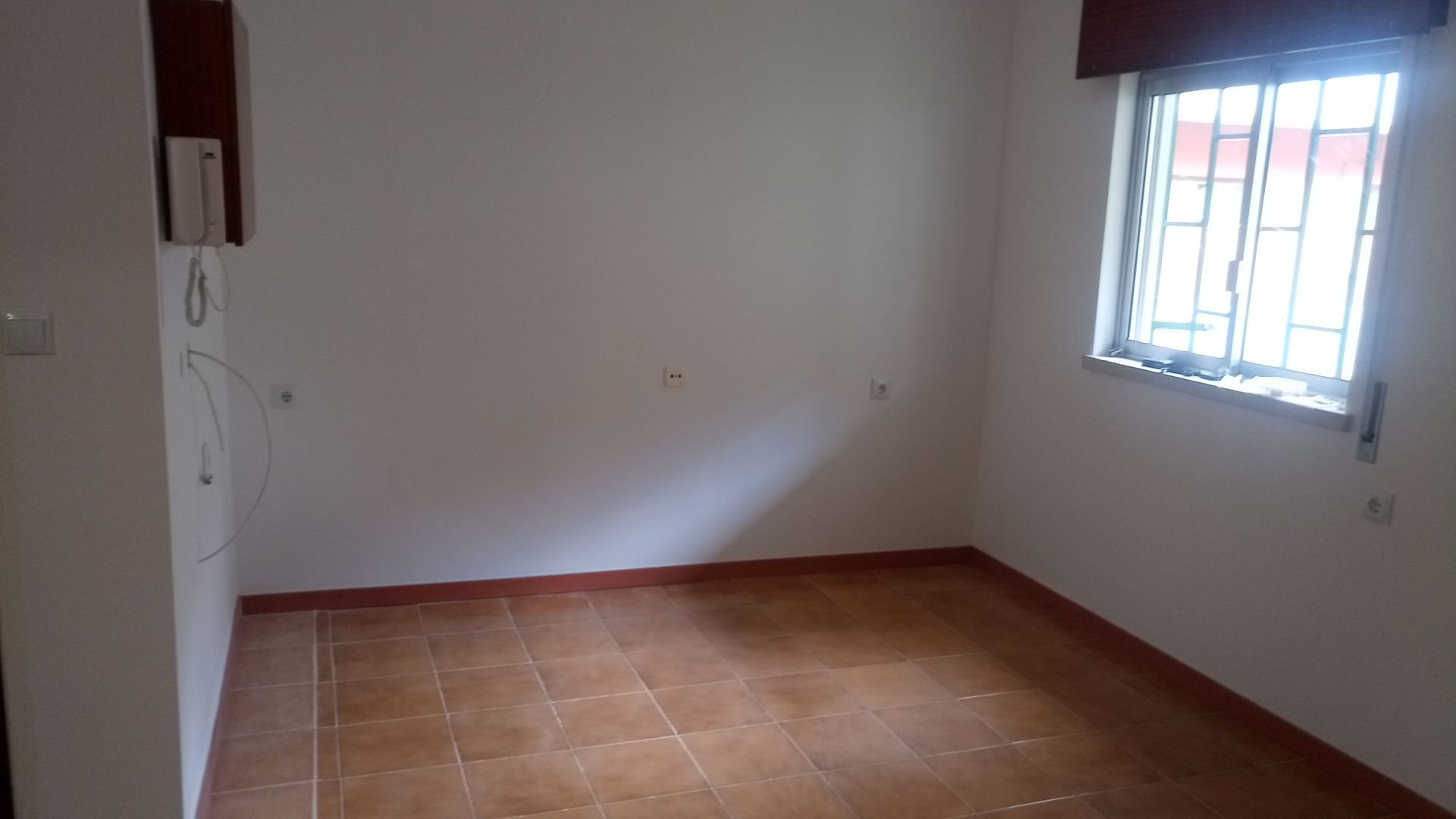 Quarto vazio com janela, piso de azulejo marrom e paredes brancas.