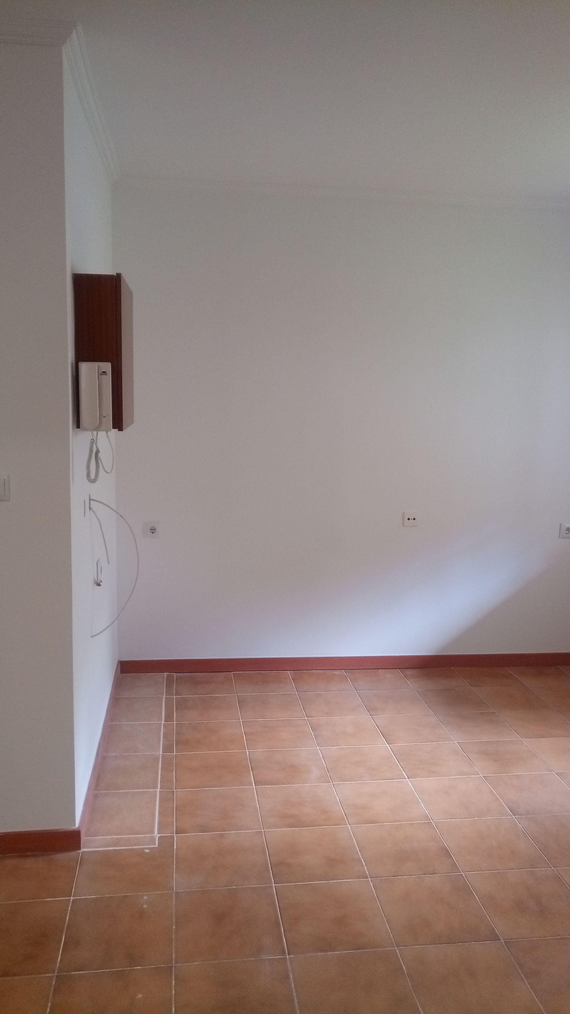 Quarto vazio com piso de azulejos marrons, paredes brancas e um pequeno armário de madeira.