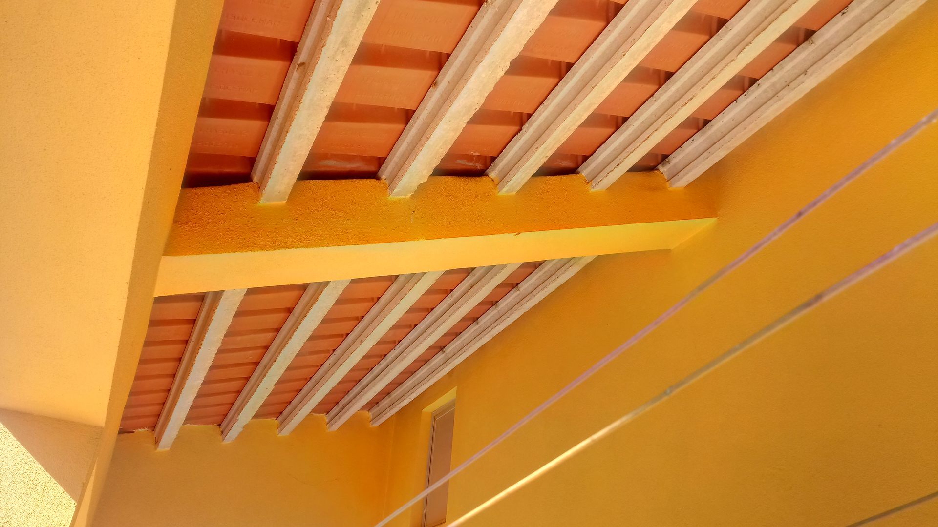 Teto com azulejos laranja e vigas brancas, visto de baixo, contra uma parede amarela.