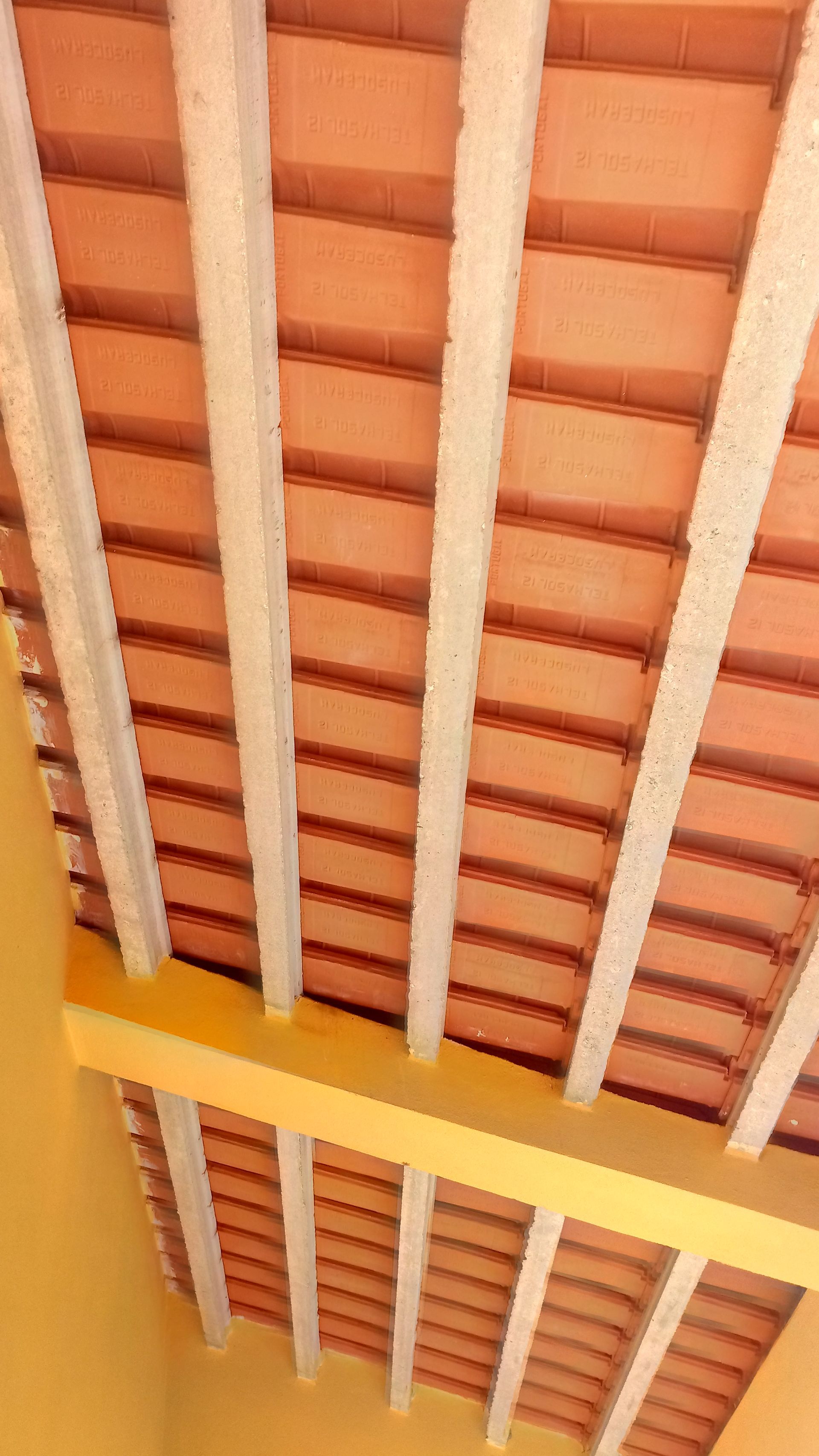 Vista de um teto com telhas de terracota, vigas brancas e uma viga de sustentação amarela.