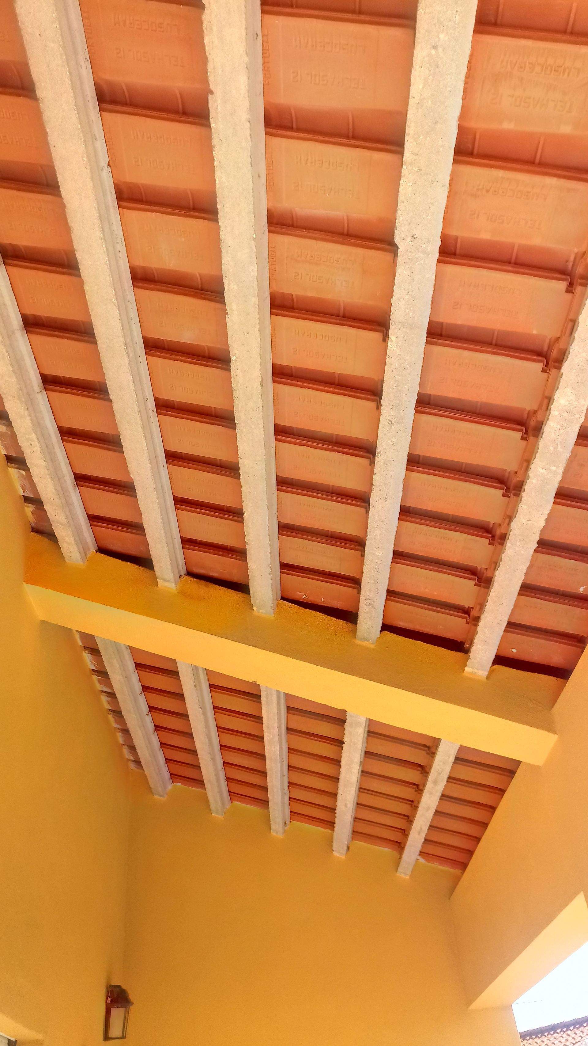 Teto com vigas aparentes e azulejos laranja, sustentado por vigas brancas e uma viga de suporte amarela, visto de baixo.