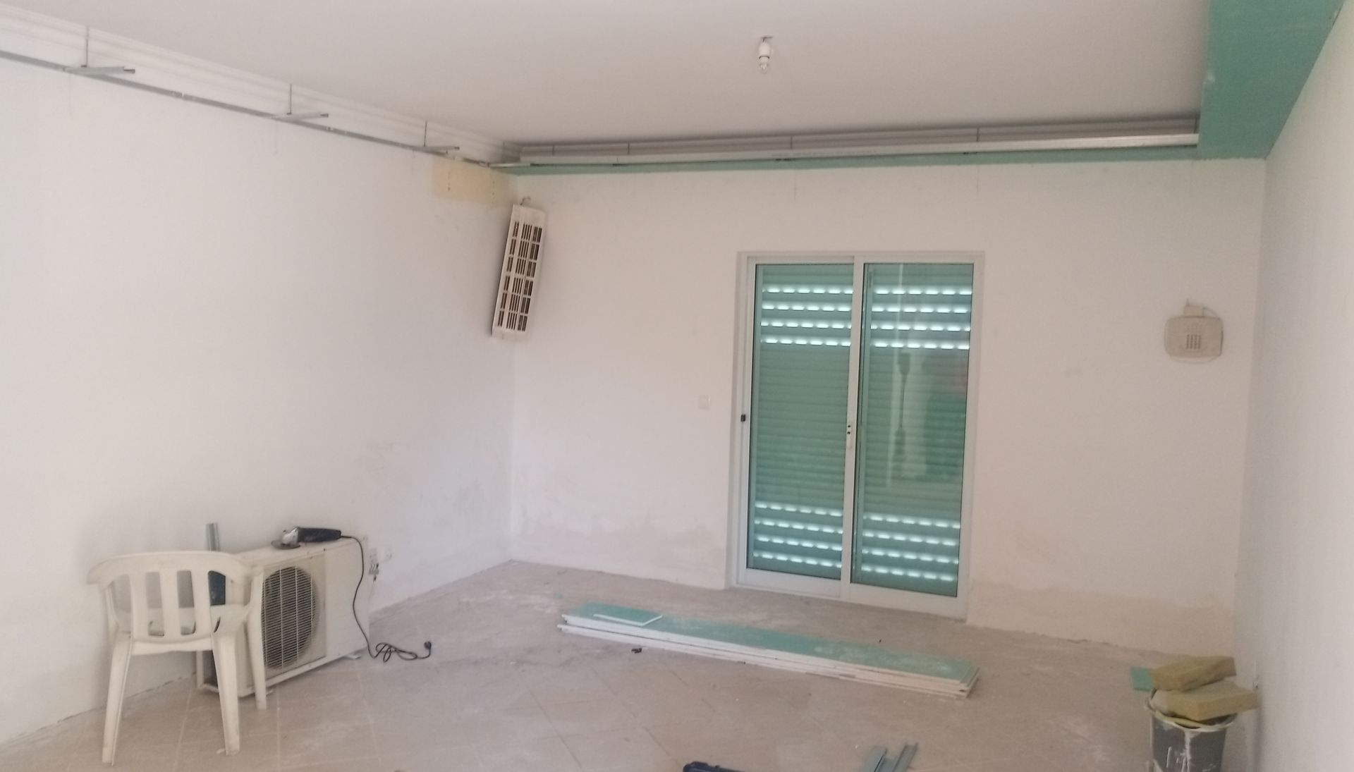 Sala vazia com paredes brancas, uma porta de vidro deslizante com persianas verdes e materiais de construção.