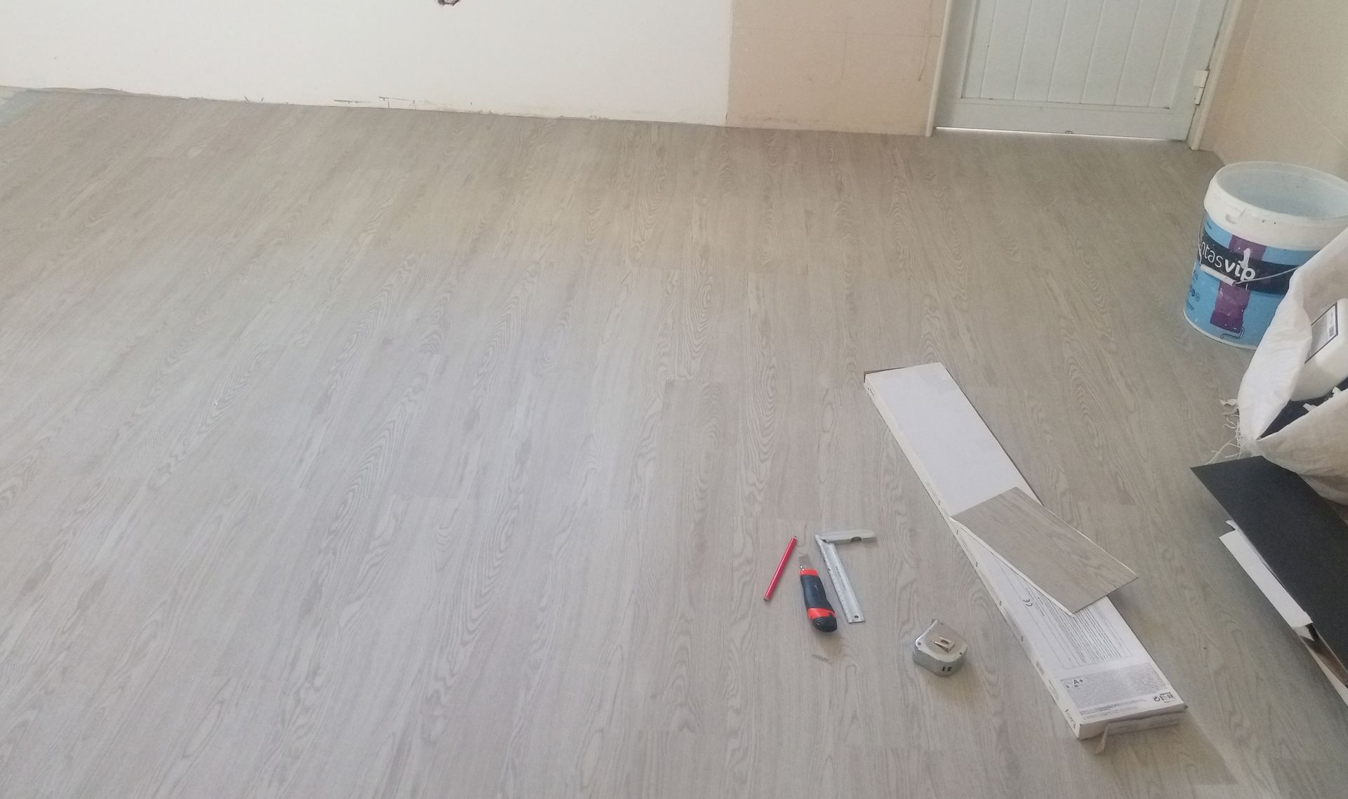 Instalando piso imitando madeira em tom claro. Ferramentas e materiais, incluindo balde de tinta, espalhados pelo chão.