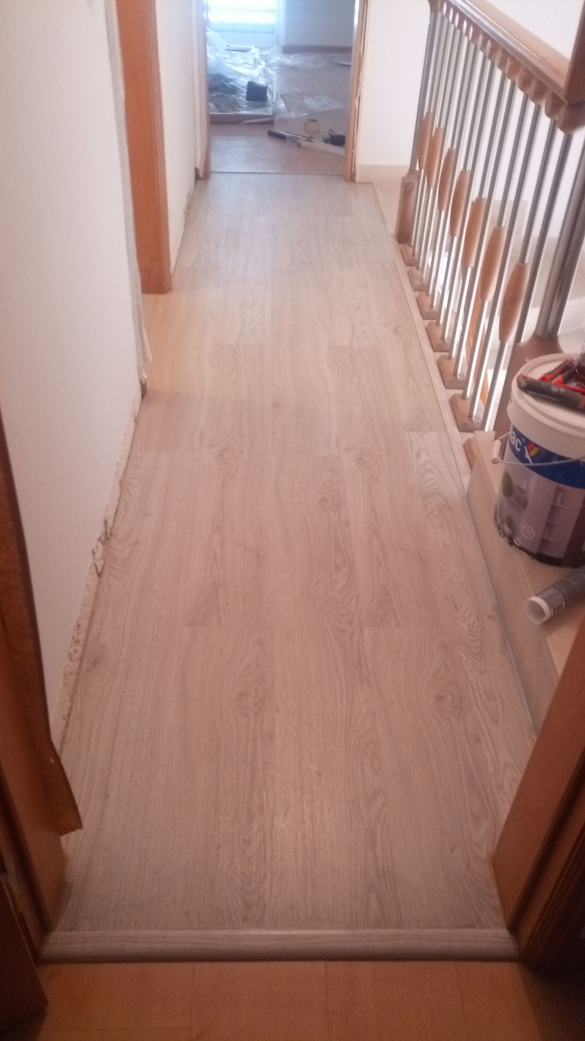 Corredor com piso de madeira clara e escada à direita. Uma lata de tinta está sobre os degraus.