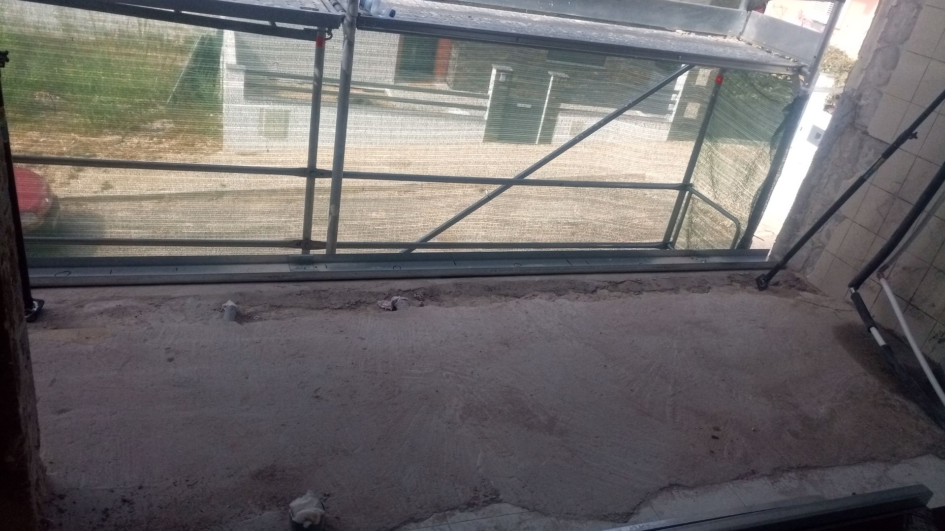 Um cômodo inacabado com uma grande janela. Andaimes visíveis do lado de fora. Chão empoeirado.