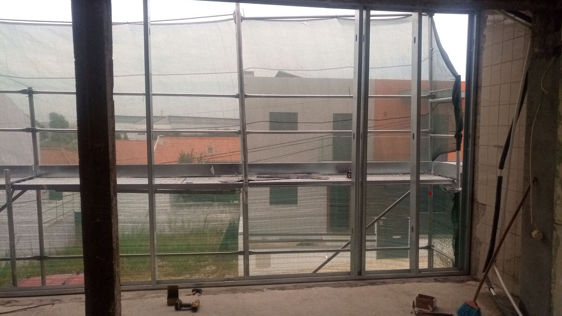Vista interna de um edifício em construção. Caixilho de janela inacabado com cobertura de tela, com vista para edifícios e andaimes.