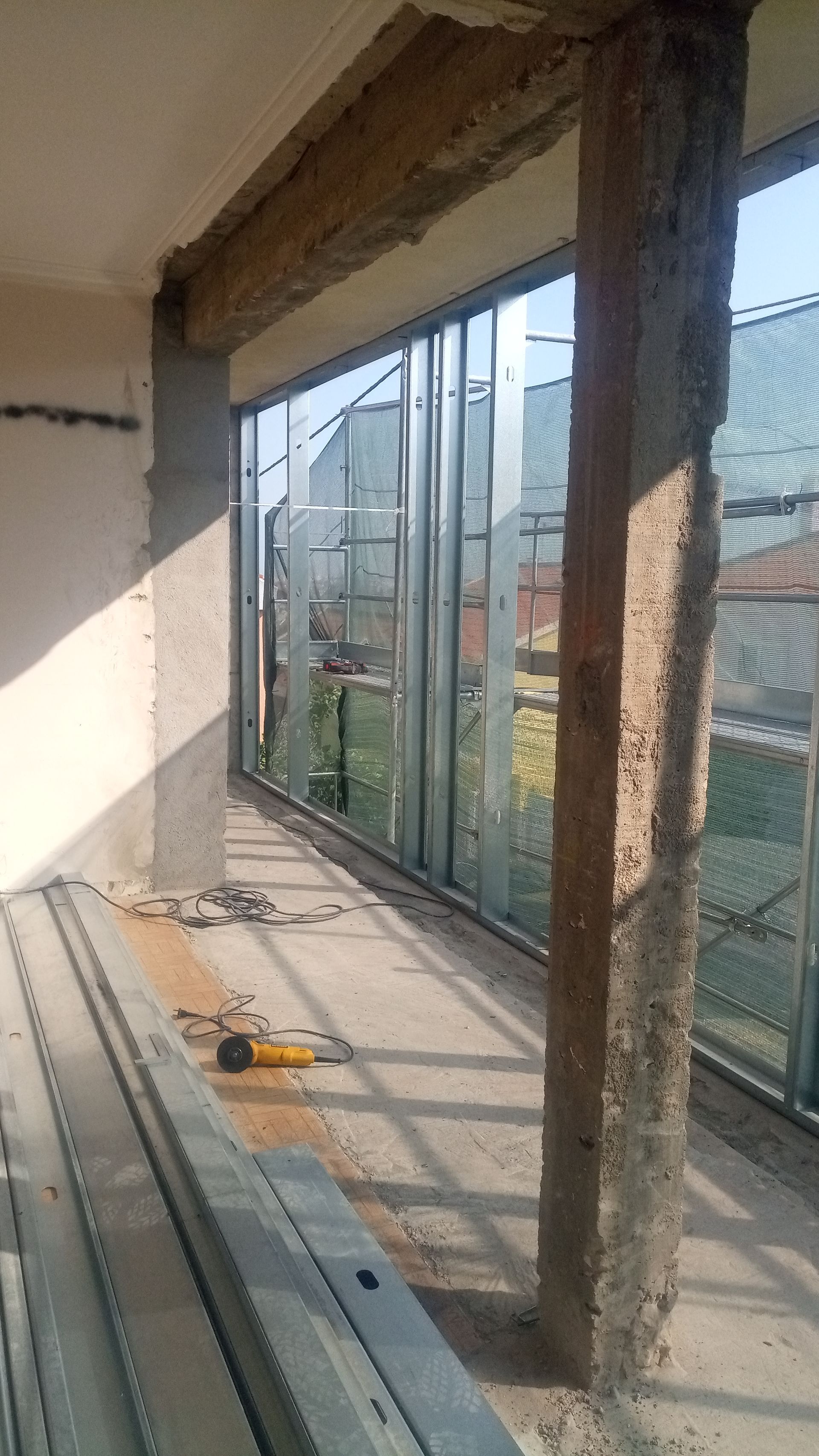 Canteiro de obras: parede com estrutura metálica em construção, vigas de concreto para sustentação, ferramentas de trabalho no chão.