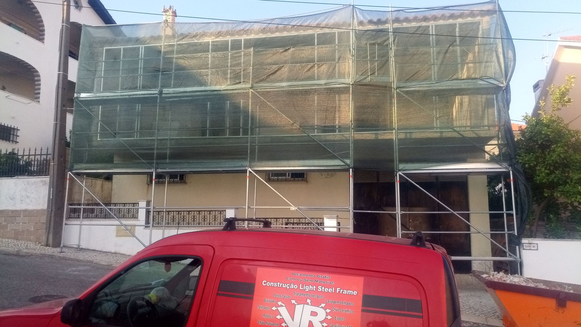 Casa em reforma, andaimes cobertos com tela verde. Uma van vermelha estacionada em frente.