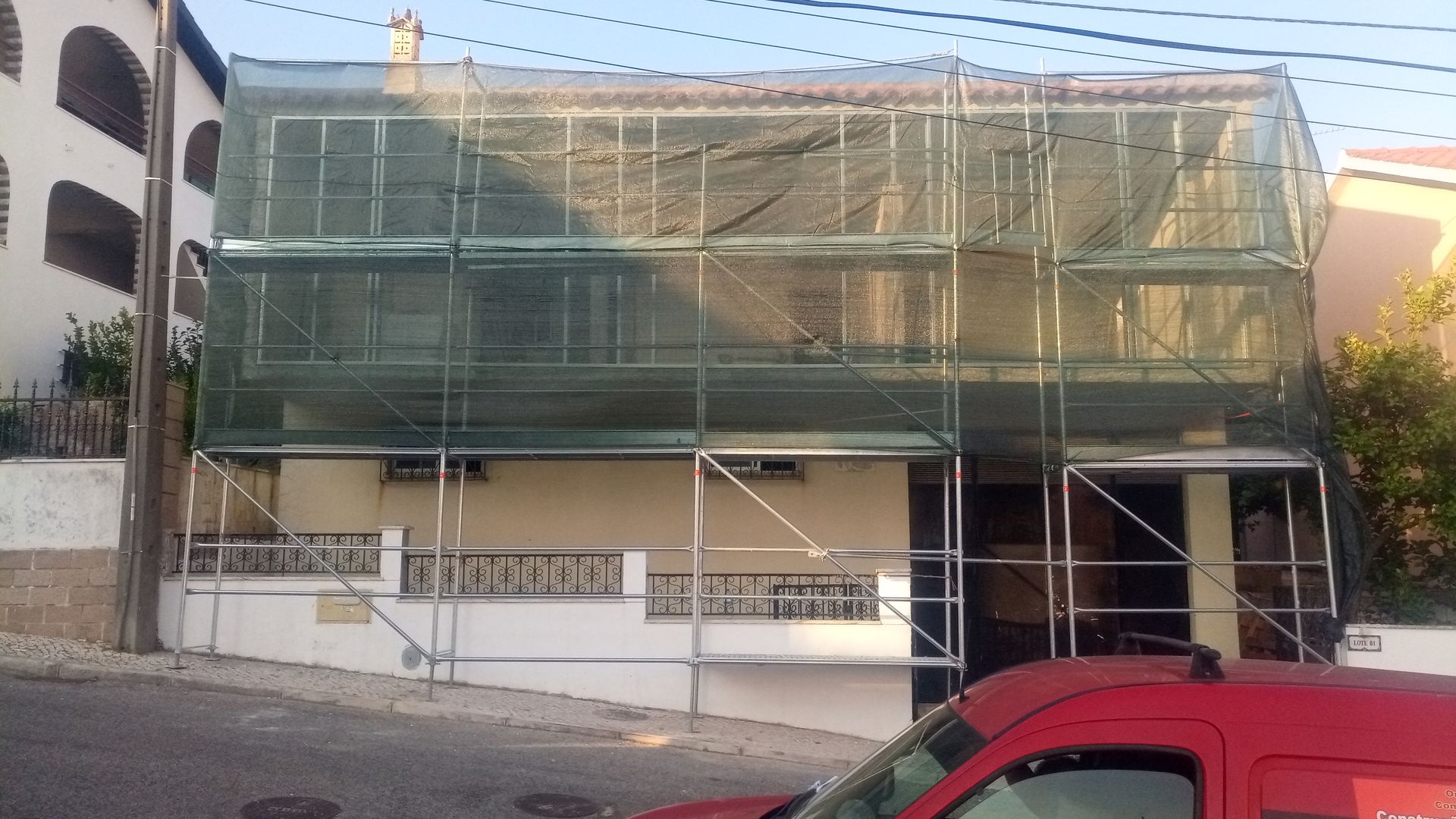 Casa coberta por redes verdes e andaimes, em reforma, em rua inclinada; um carro vermelho está estacionado em primeiro plano.