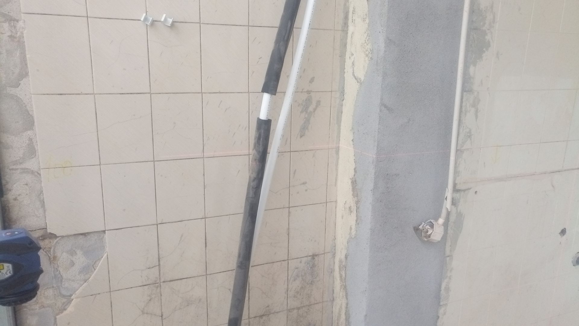 Canteiro de obras: Parede revestida com azulejos parcialmente demolida, coluna de concreto exposta e ferramentas encostadas na parede.