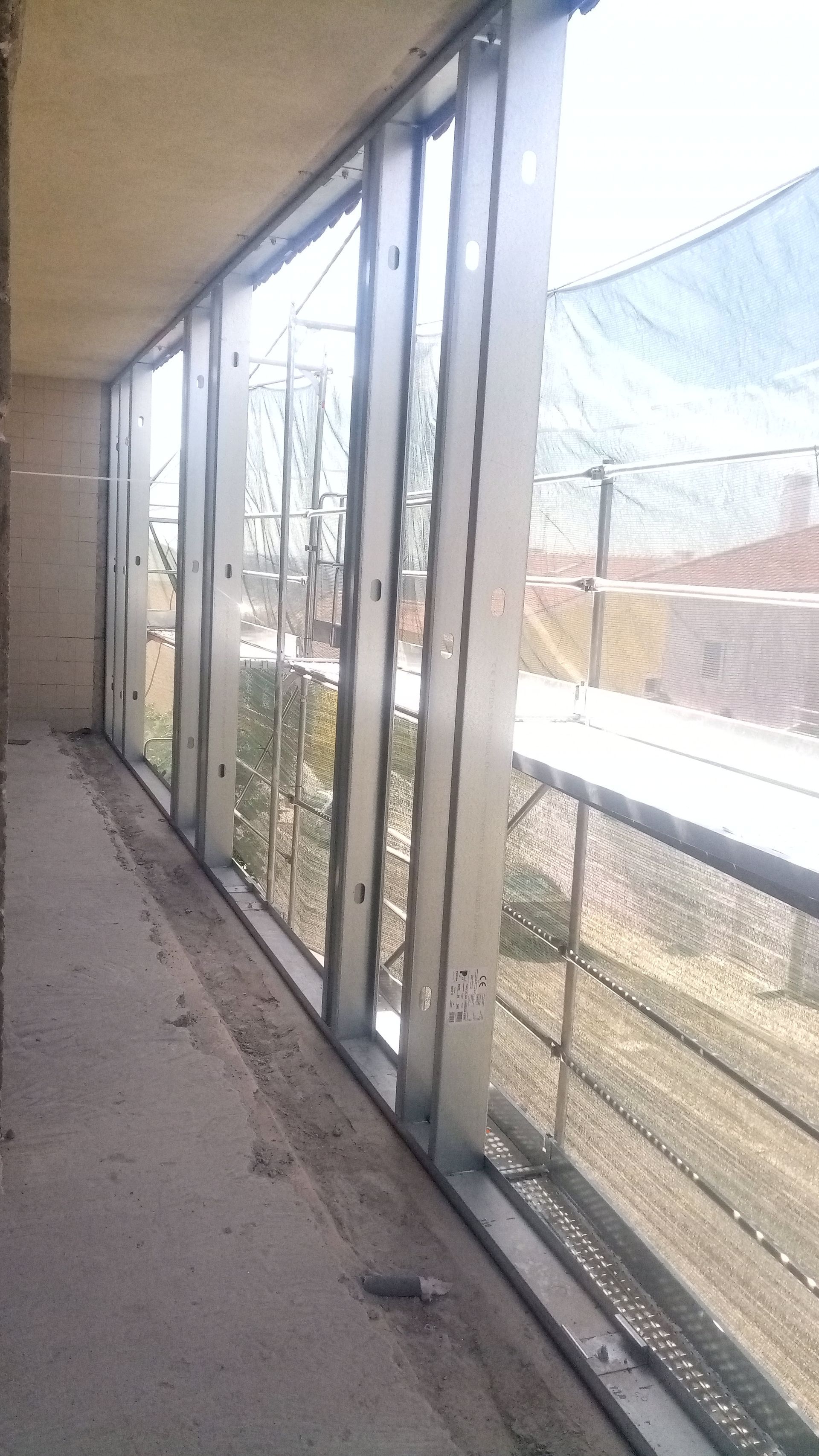 Uma estrutura de janela parcialmente construída, em metal e vidro, com vista para um exterior luminoso com gradeamento.