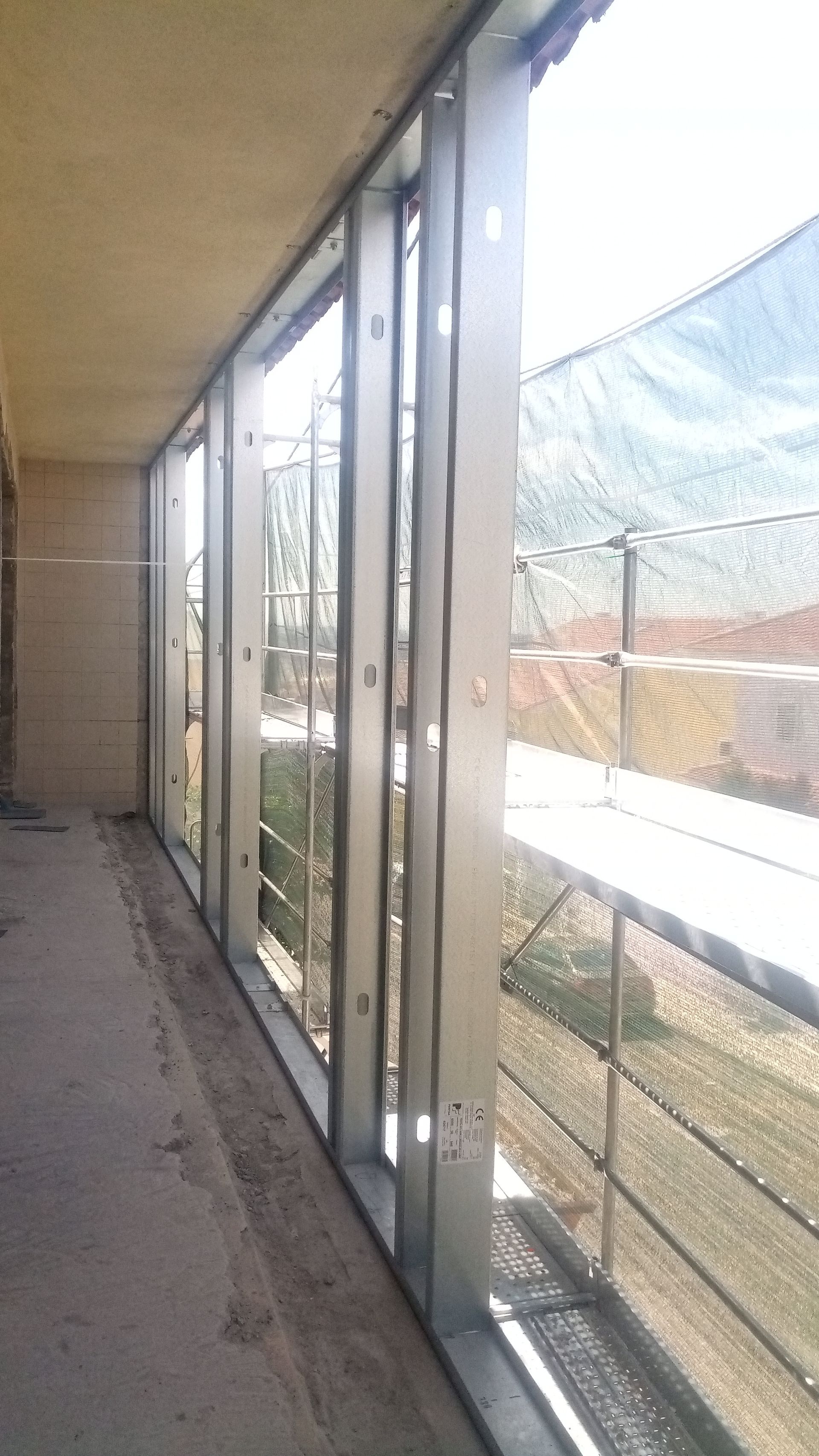 Vista interior de um edifício inacabado com caixilhos de metal nas janelas, com vista para um pátio com andaimes e redes de proteção.